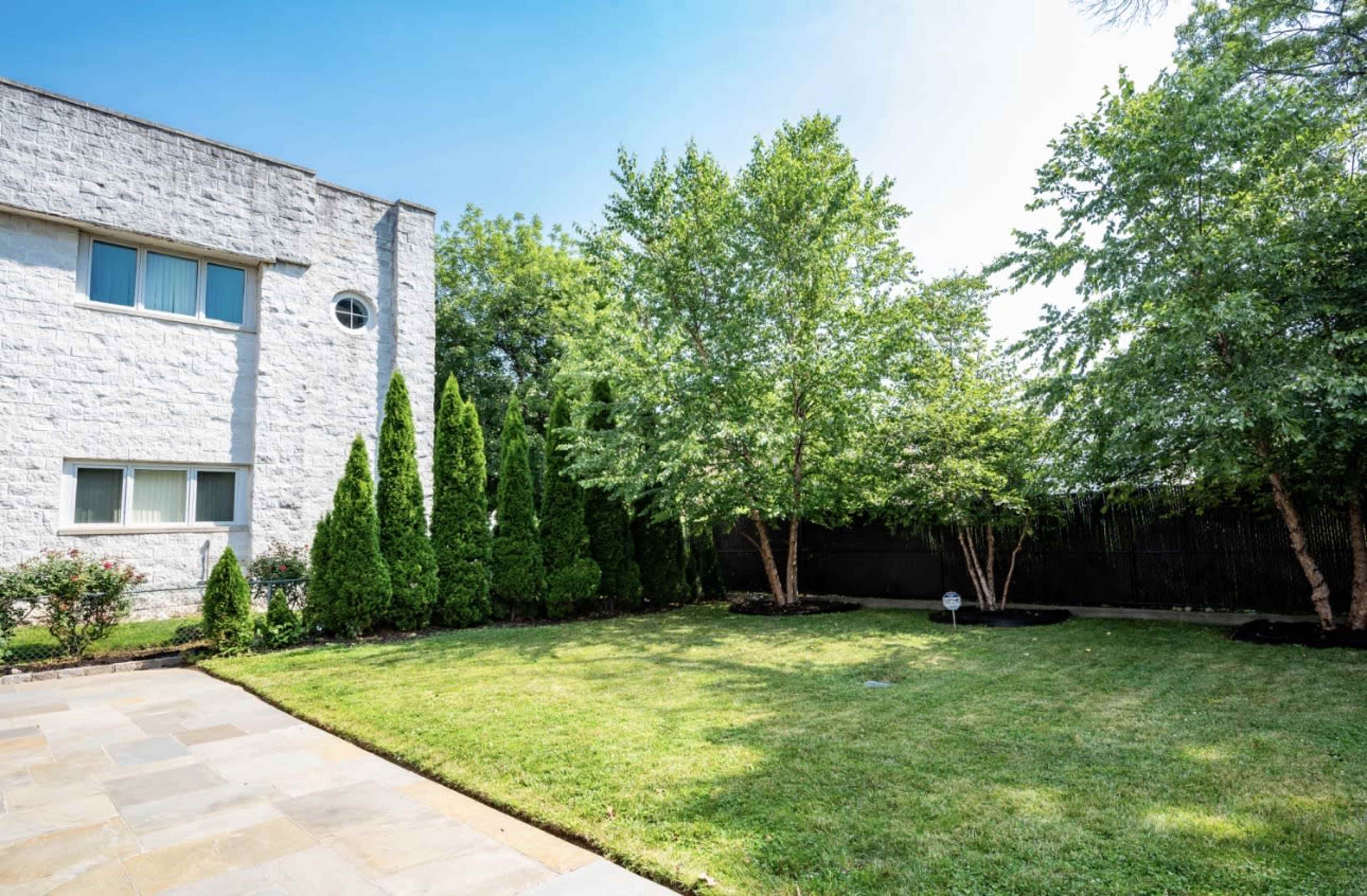The image shows a neatly maintained backyard with a green lawn, several small trees, and a stone patio adjacent to a white building.