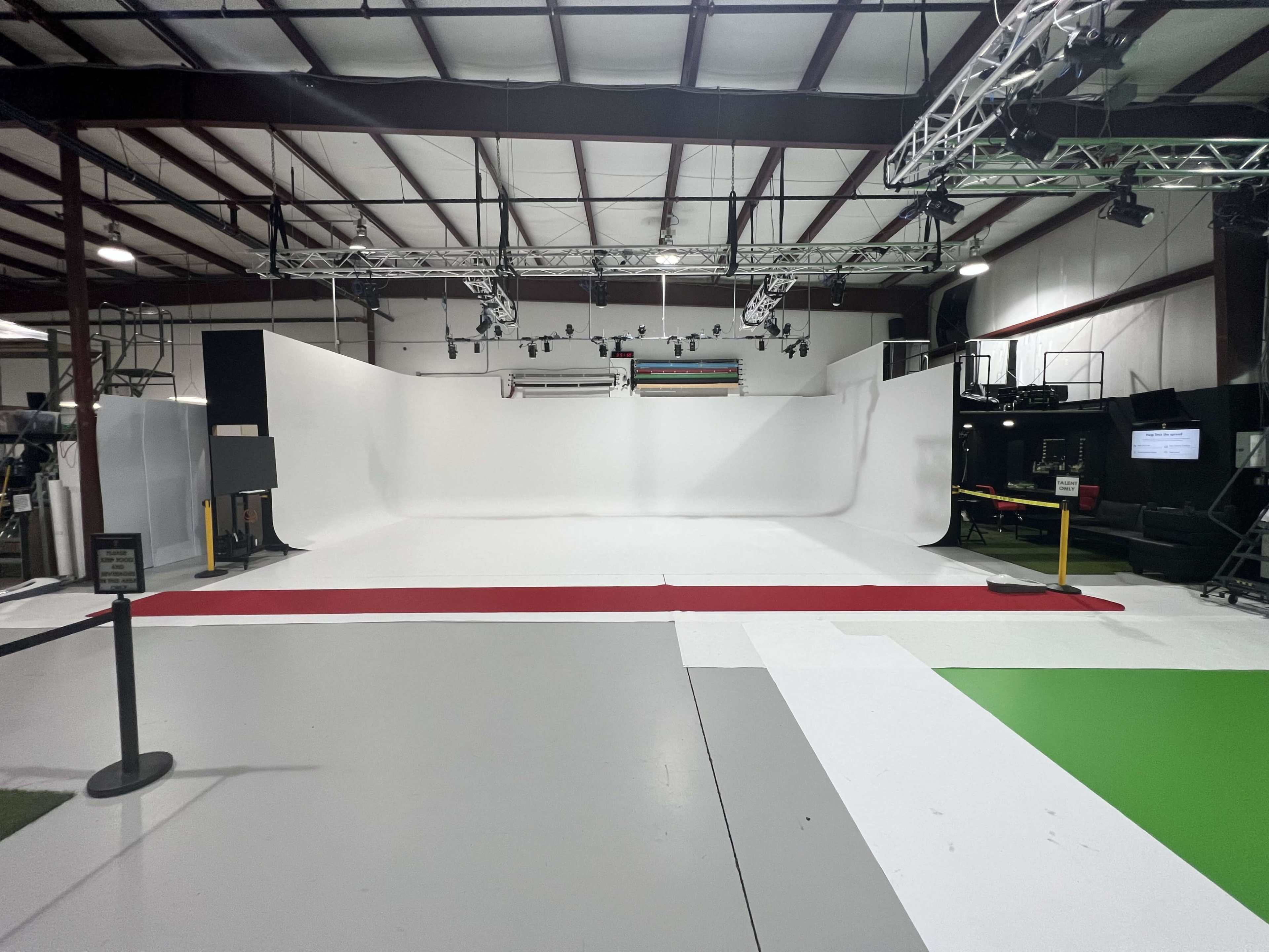 The image shows a spacious studio with a white backdrop and various lighting equipment, featuring a colored carpet and sections of different flooring.