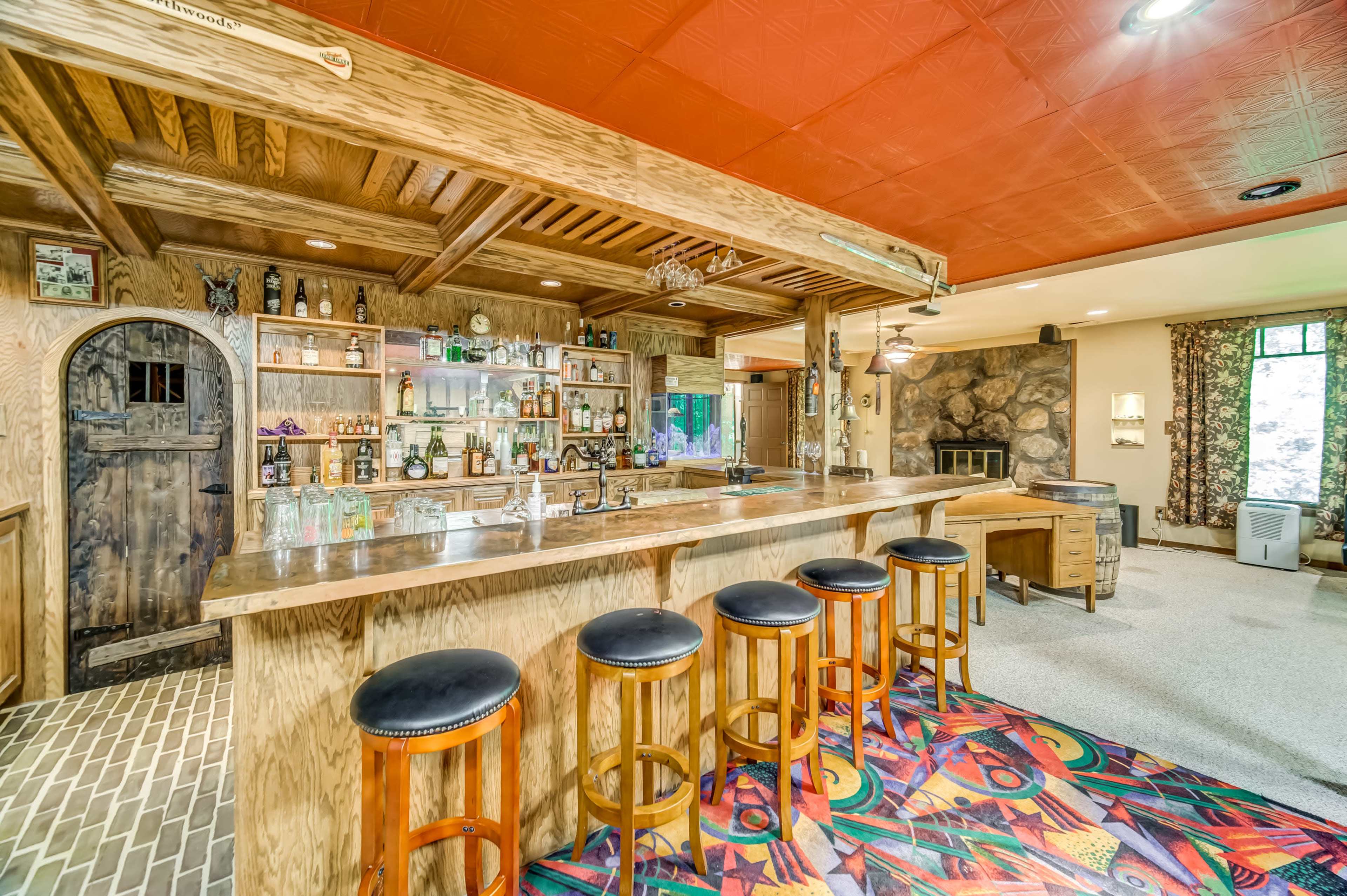 The image shows a wooden bar area with high stools, shelves of alcohol, and a stone fireplace in a lounge setting.