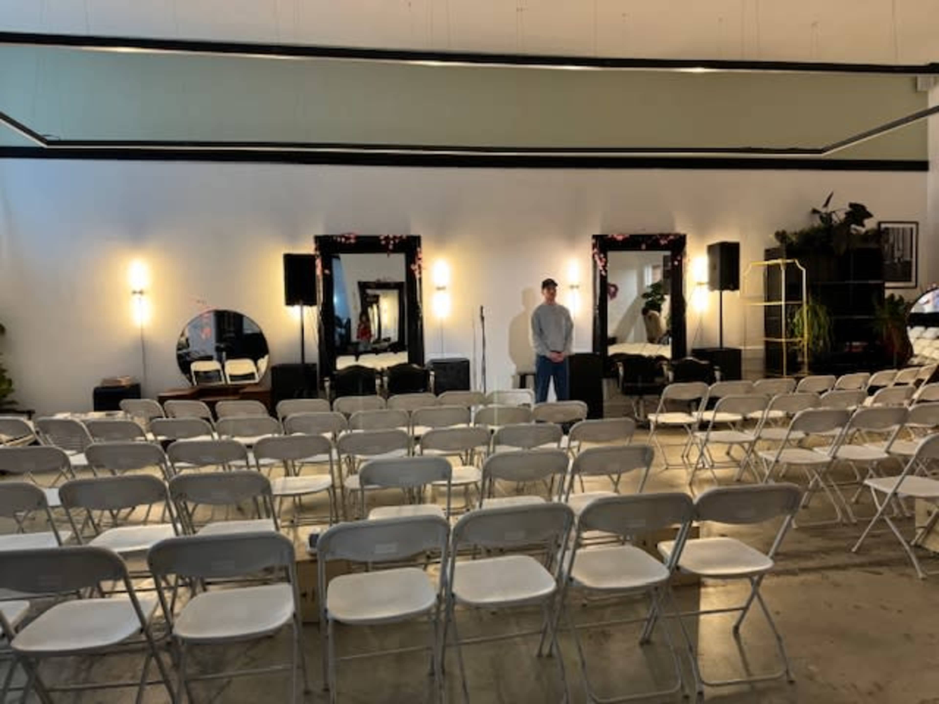 The image shows a sparsely decorated event space with rows of white folding chairs facing a stage area featuring three mirrors and a person standing in front of them.