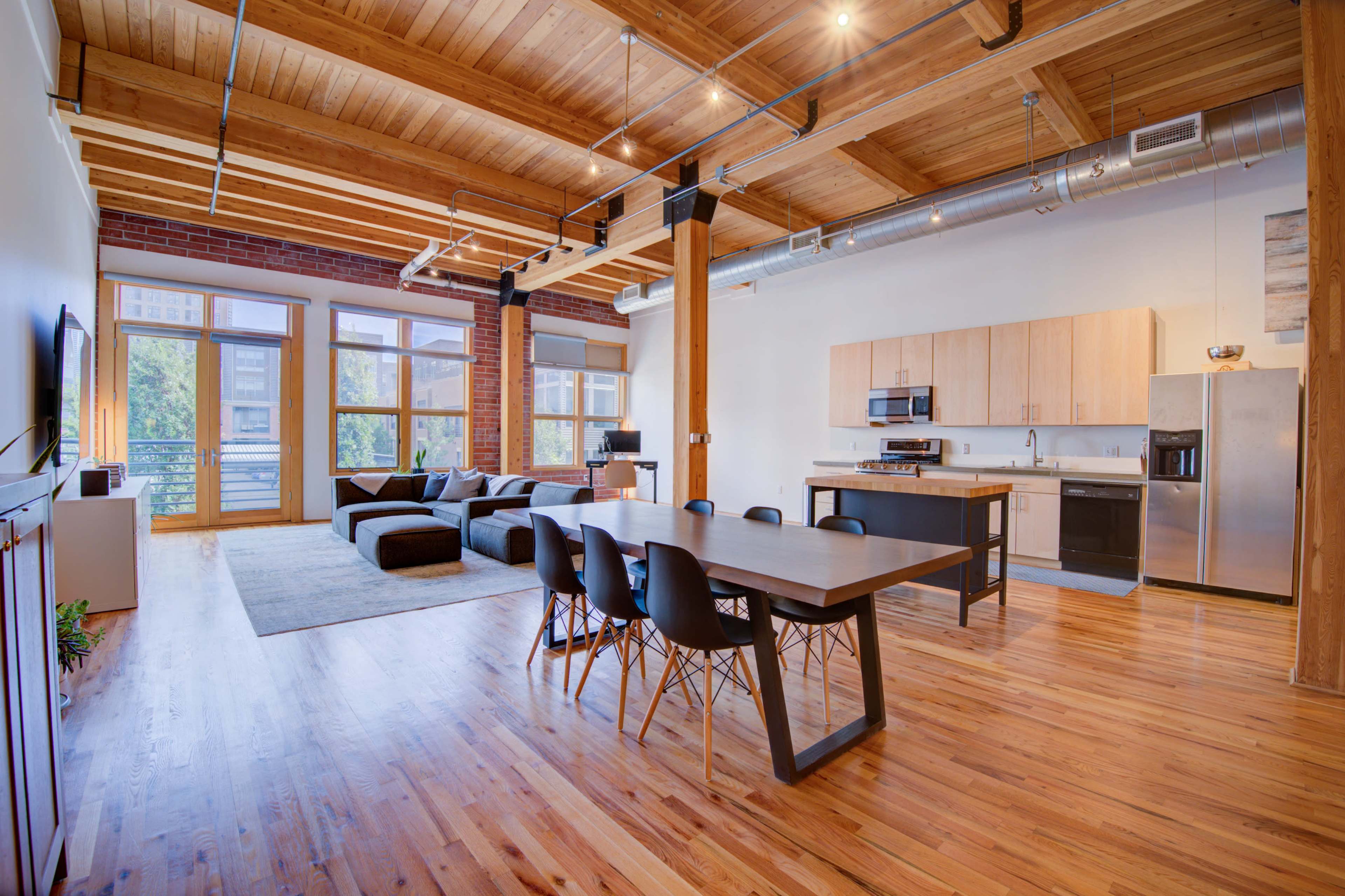 The image shows a spacious open-concept living area featuring a kitchen, dining table, and a lounge with a large sectional sofa, all under exposed wooden beams and bright windows.
