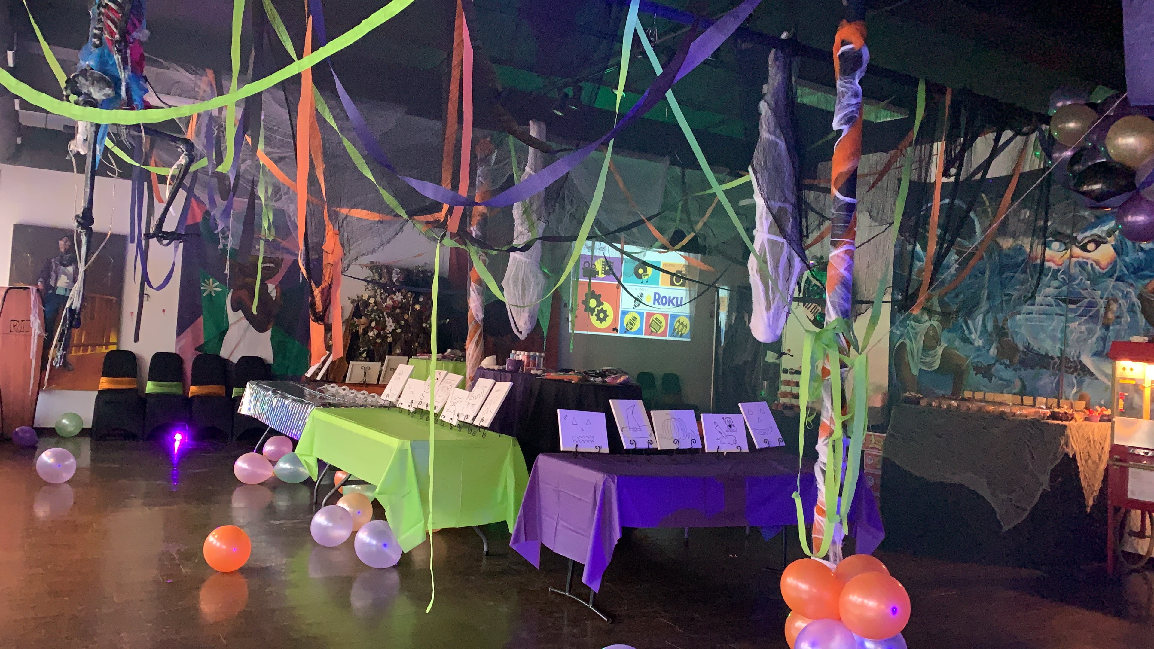 A decorated event space features colorful streamers and balloons, with tables displaying various items and a projection screen in the background.