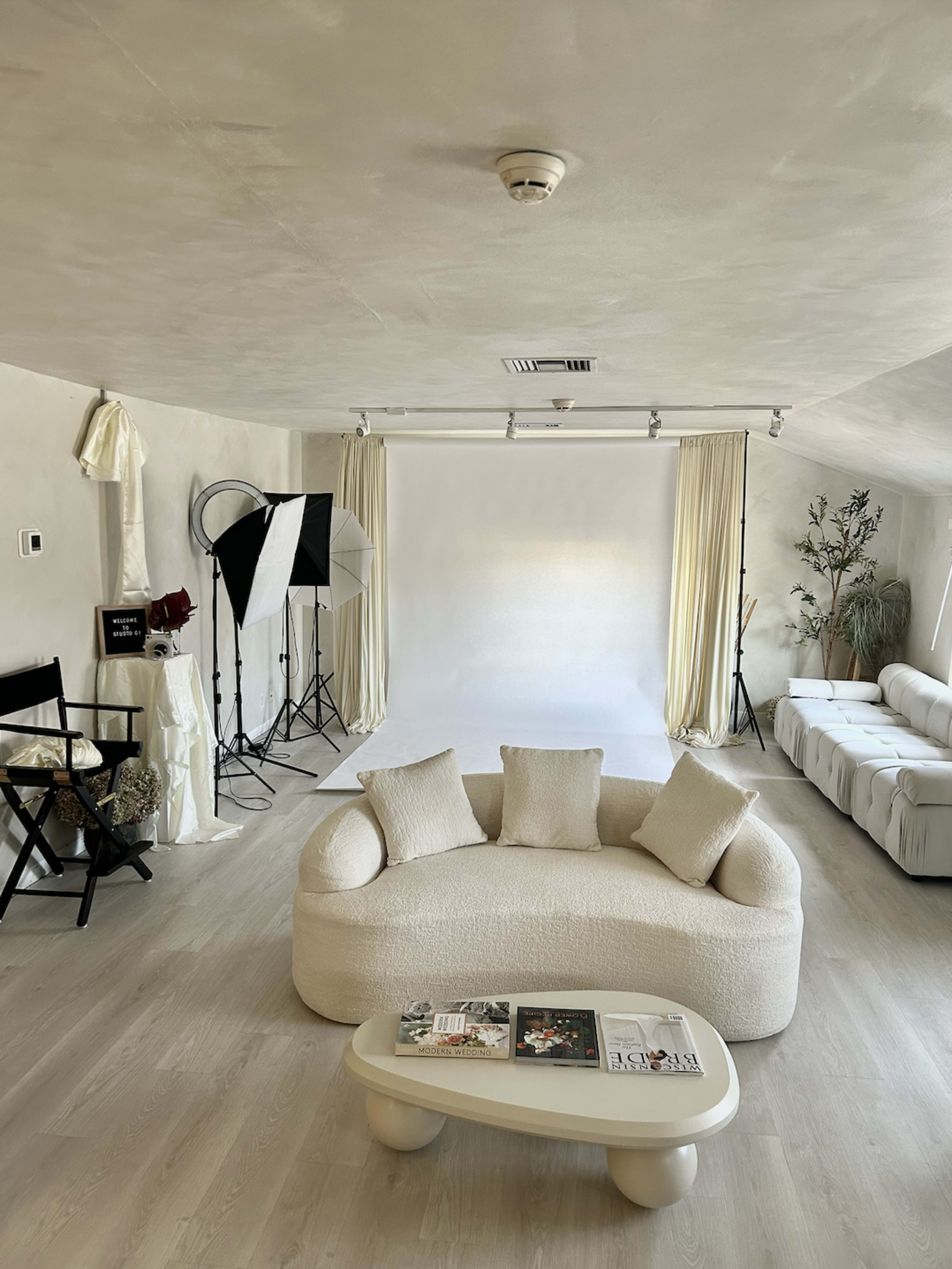 The image shows a modern photo studio featuring a circular sofa, a white backdrop, lighting equipment, and a simple coffee table with magazines.