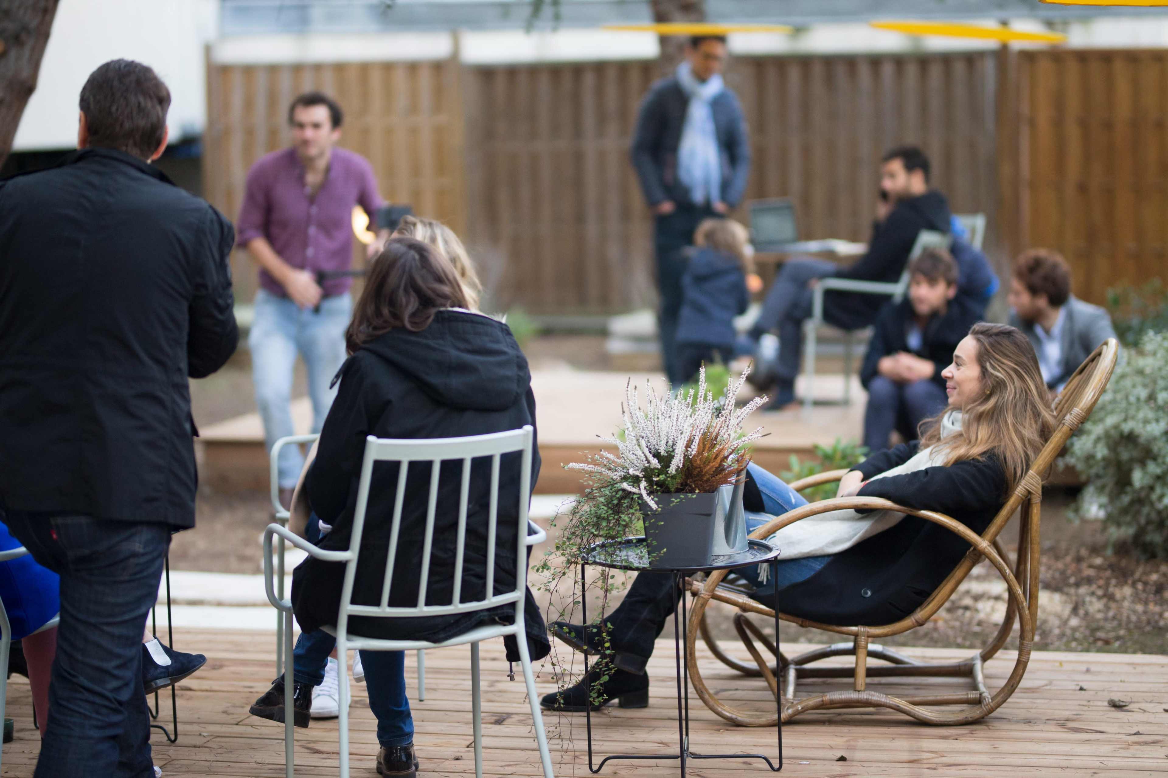 A group of people are gathered outdoors, some seated on chairs and others standing, in a casual social setting with greenery and a wooden deck.