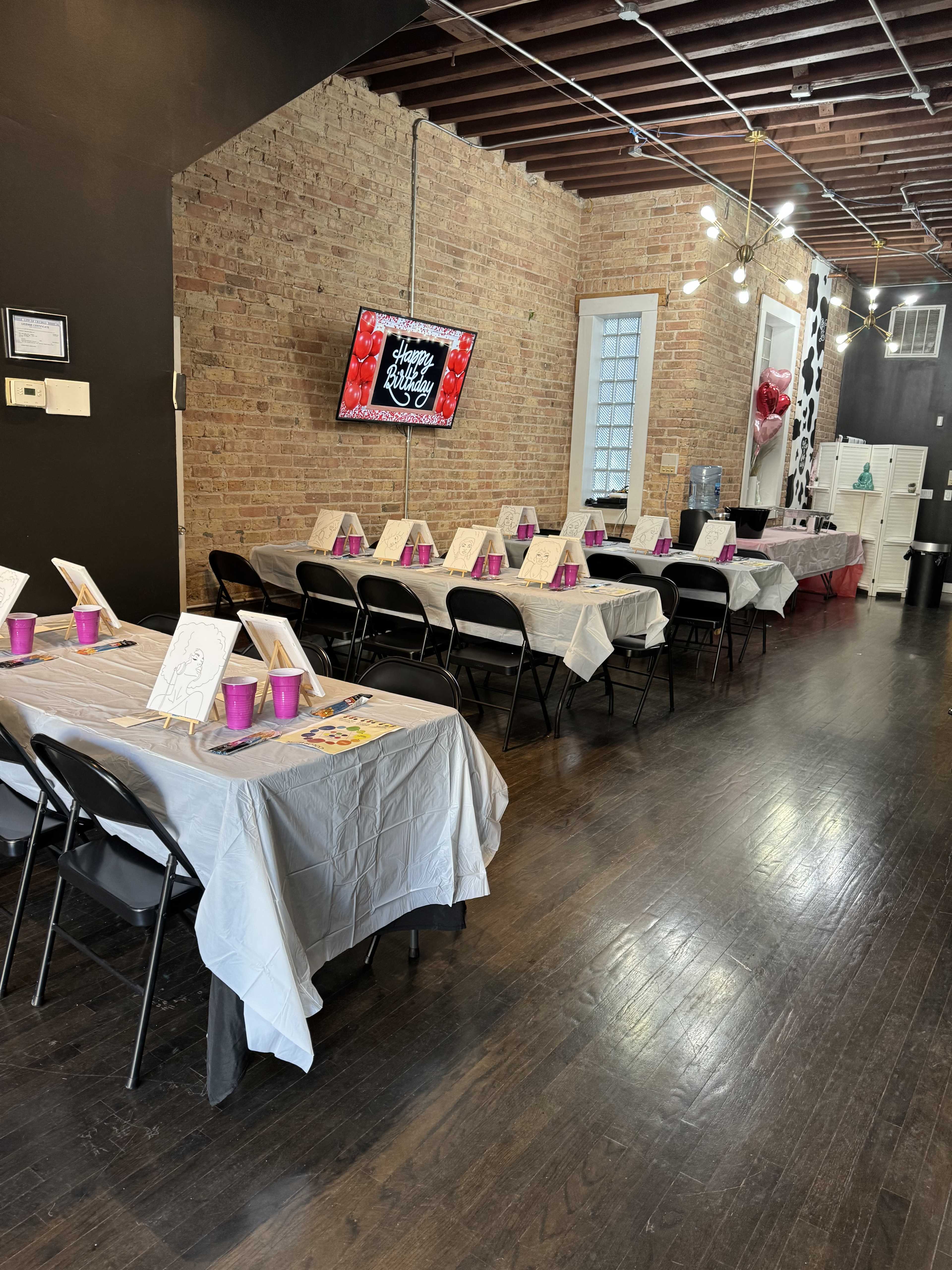 A row of tables covered with white tablecloths and decorated with pink cups in a room with brick walls and string lights.