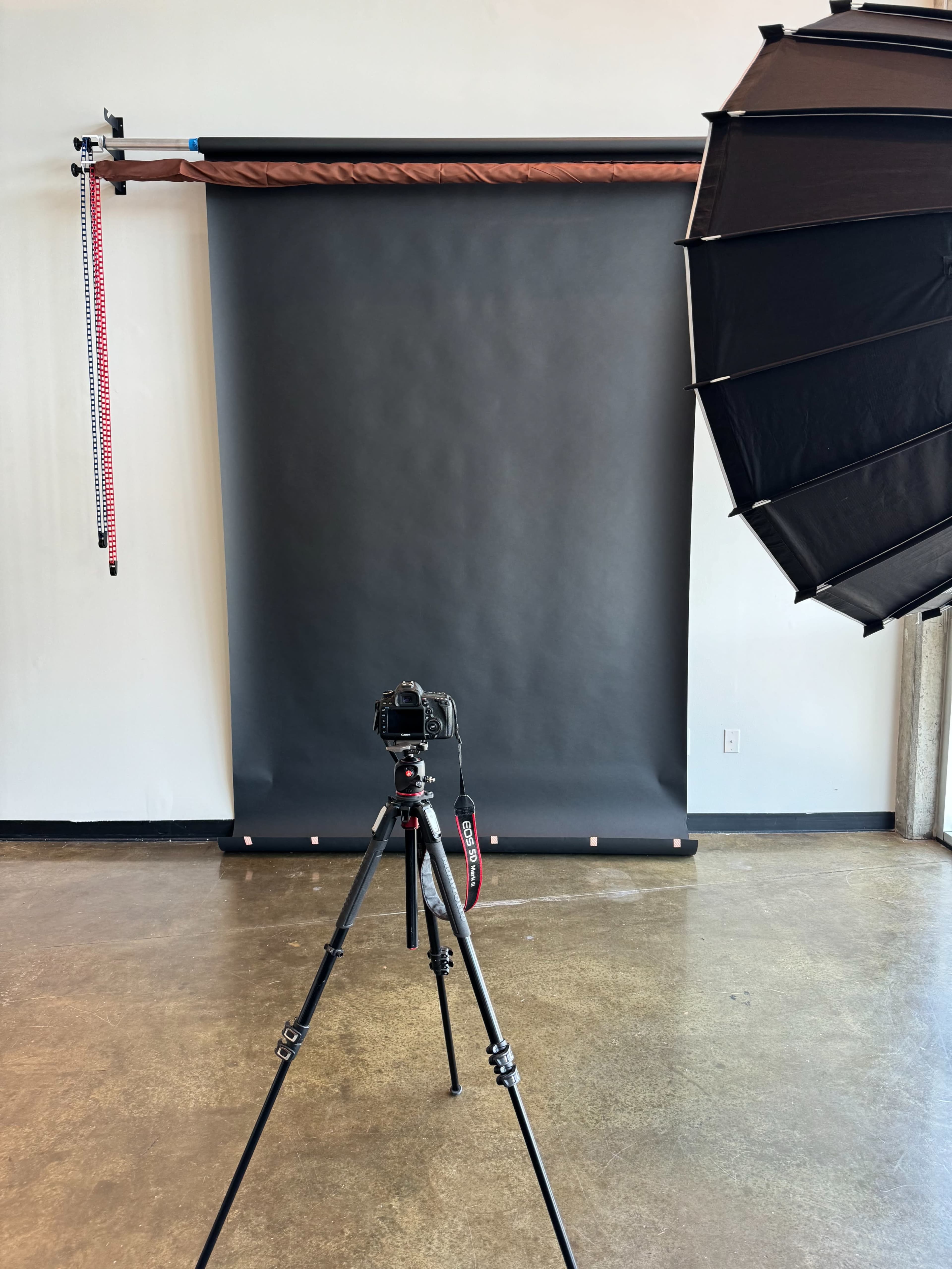 A camera mounted on a tripod is positioned in front of a dark backdrop with a softbox lighting setup to the side.