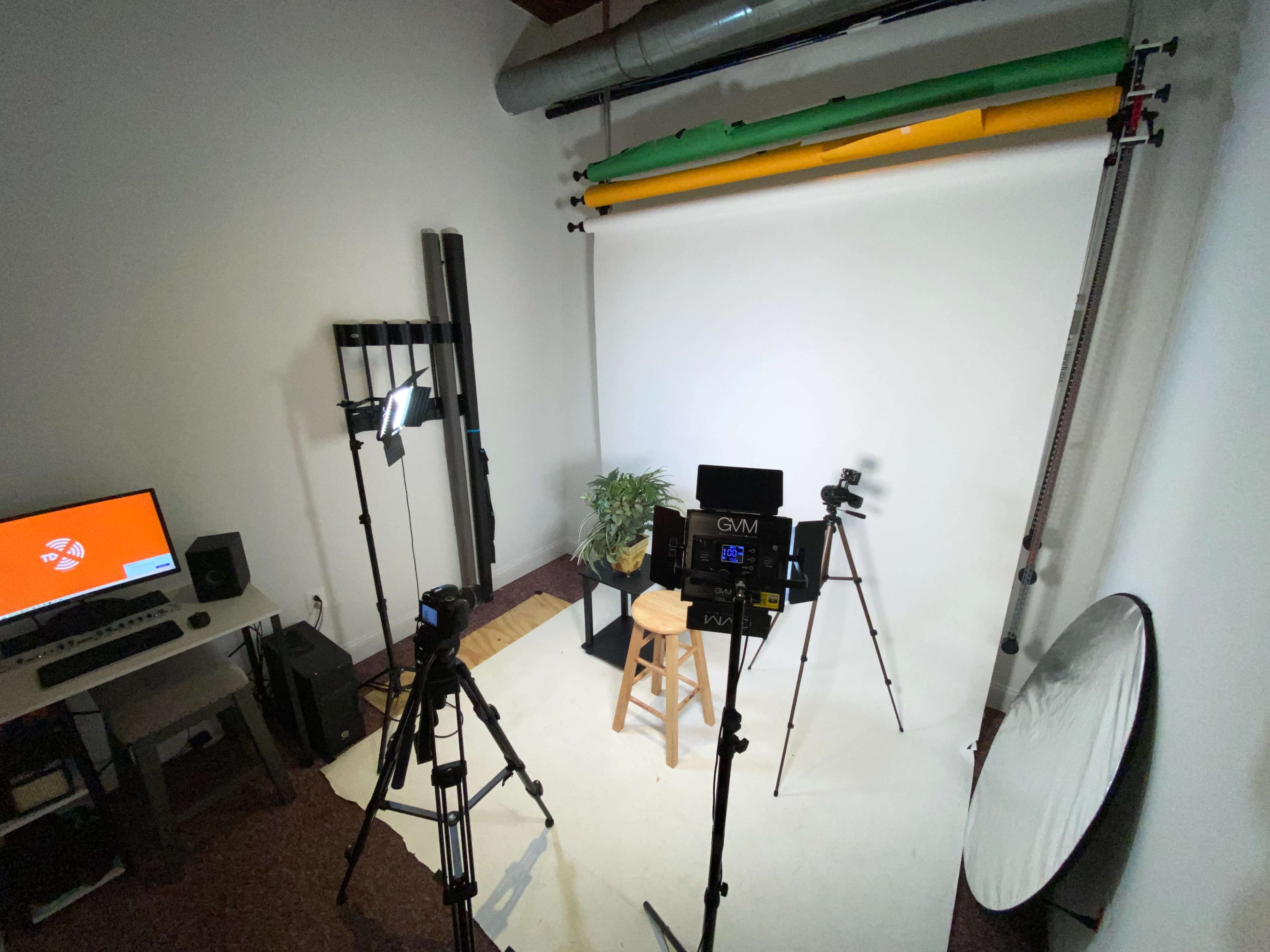 The image shows a photography studio setup with a white backdrop, studio lights, cameras on tripods, and a stool for a subject.