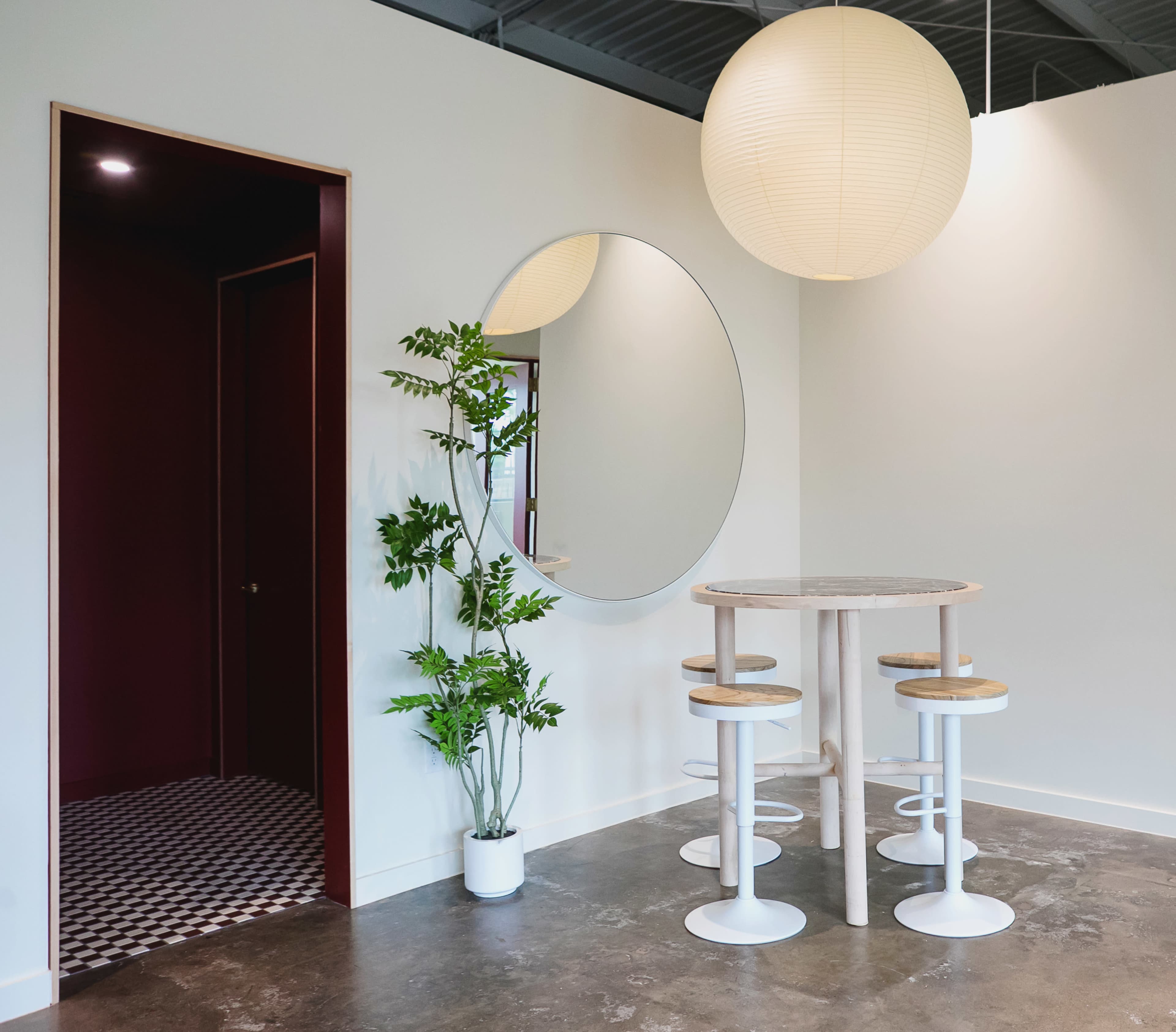 The image shows a modern interior space featuring a round mirror, a small circular table with stools, a potted plant, and a doorway leading to another room.