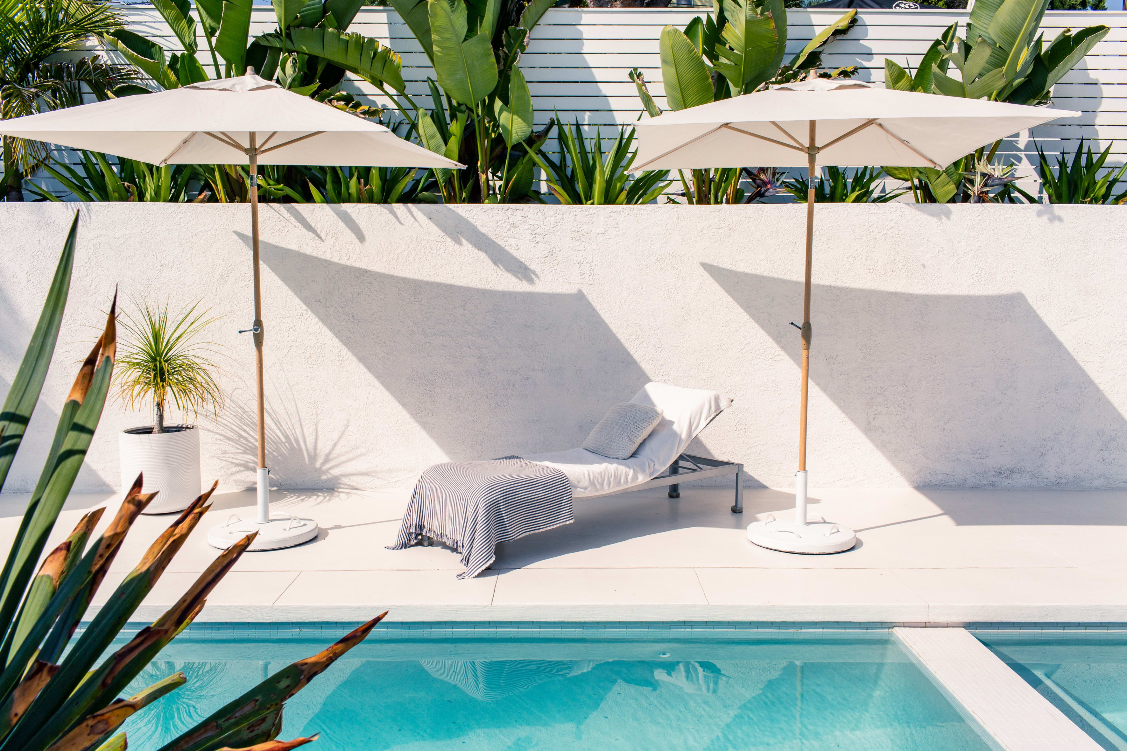 A sun lounger with a gray and white towel is placed beside a swimming pool, shaded by two white umbrellas, with lush greenery in the background.