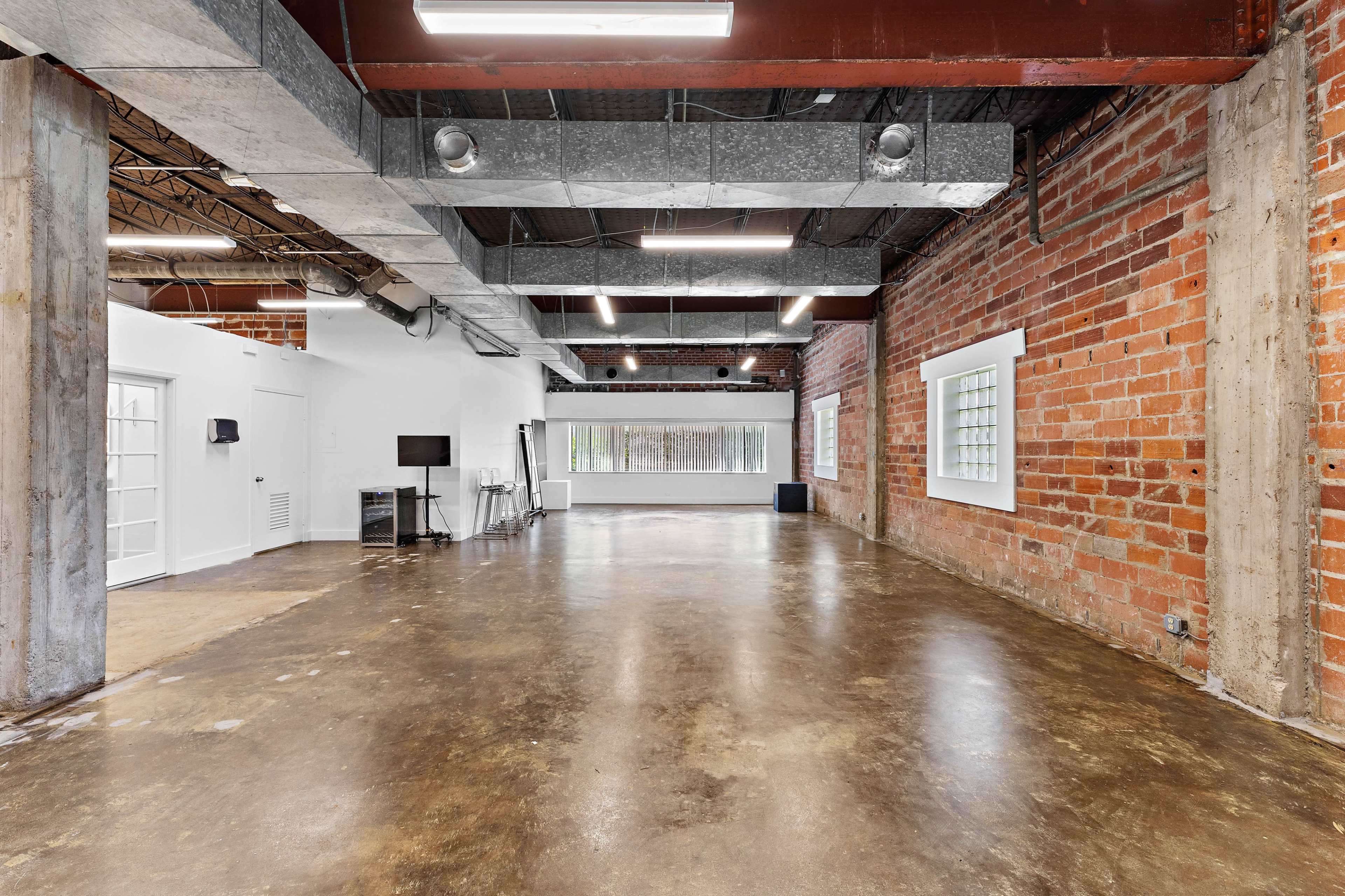 The image shows an open, industrial-style interior space with exposed brick walls, polished concrete flooring, and large windows allowing natural light.