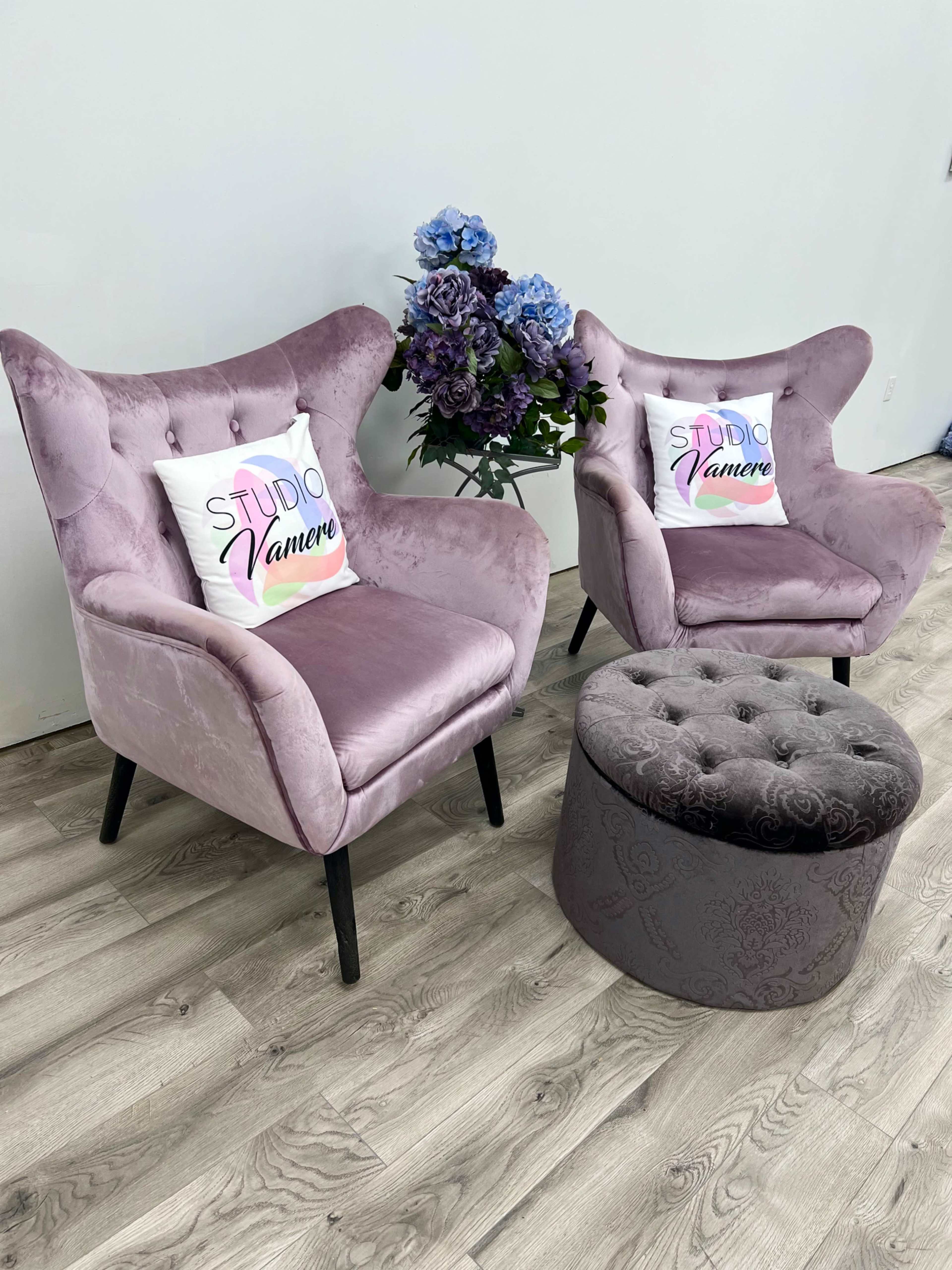 Two pink armchairs with decorative pillows positioned next to a round upholstered ottoman, set against a light-colored wall and adorned with a bouquet of blue flowers.