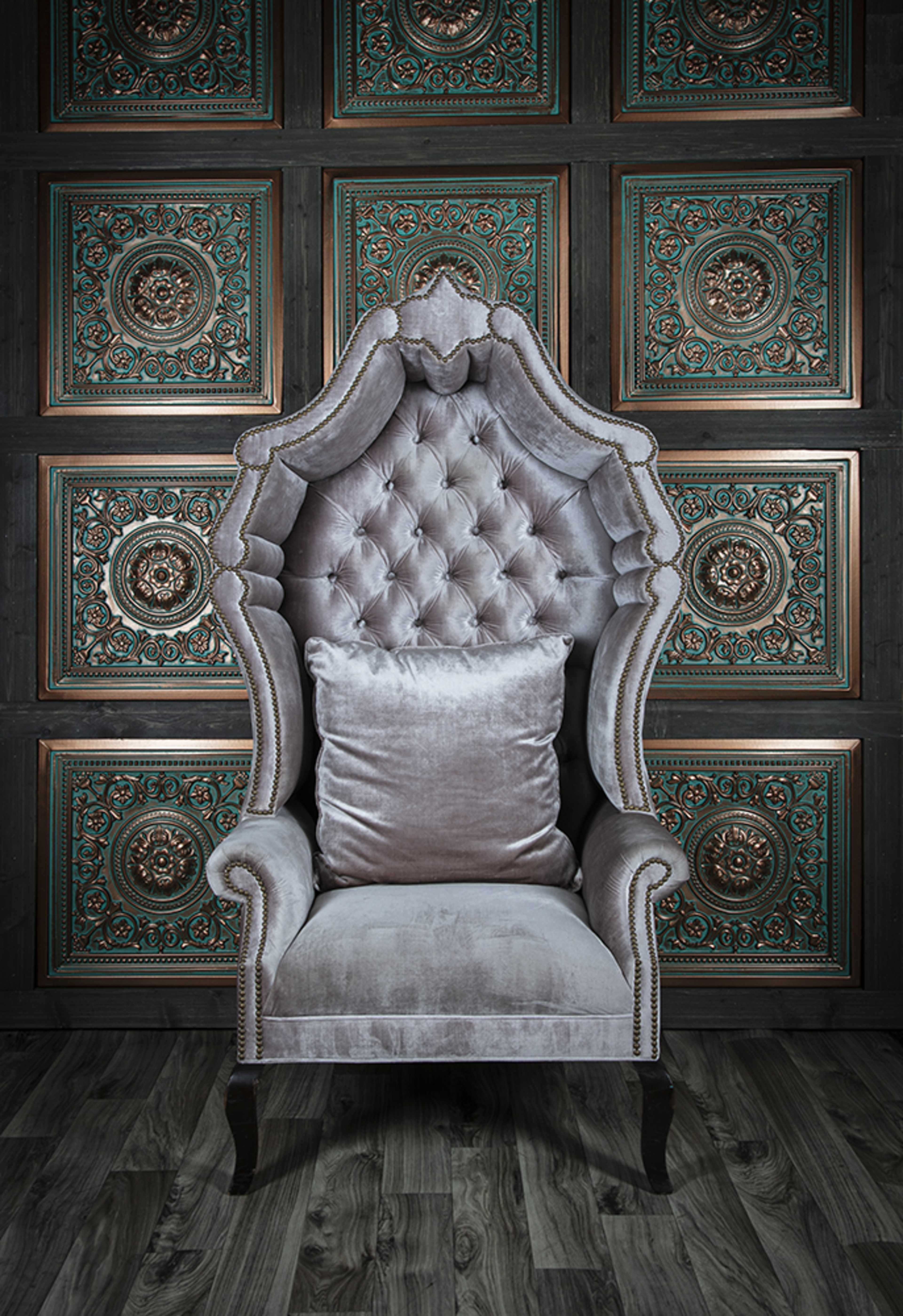 The image features an ornate, gray velvet tufted armchair against a dark wood-paneled wall with intricate teal and gold detailing.
