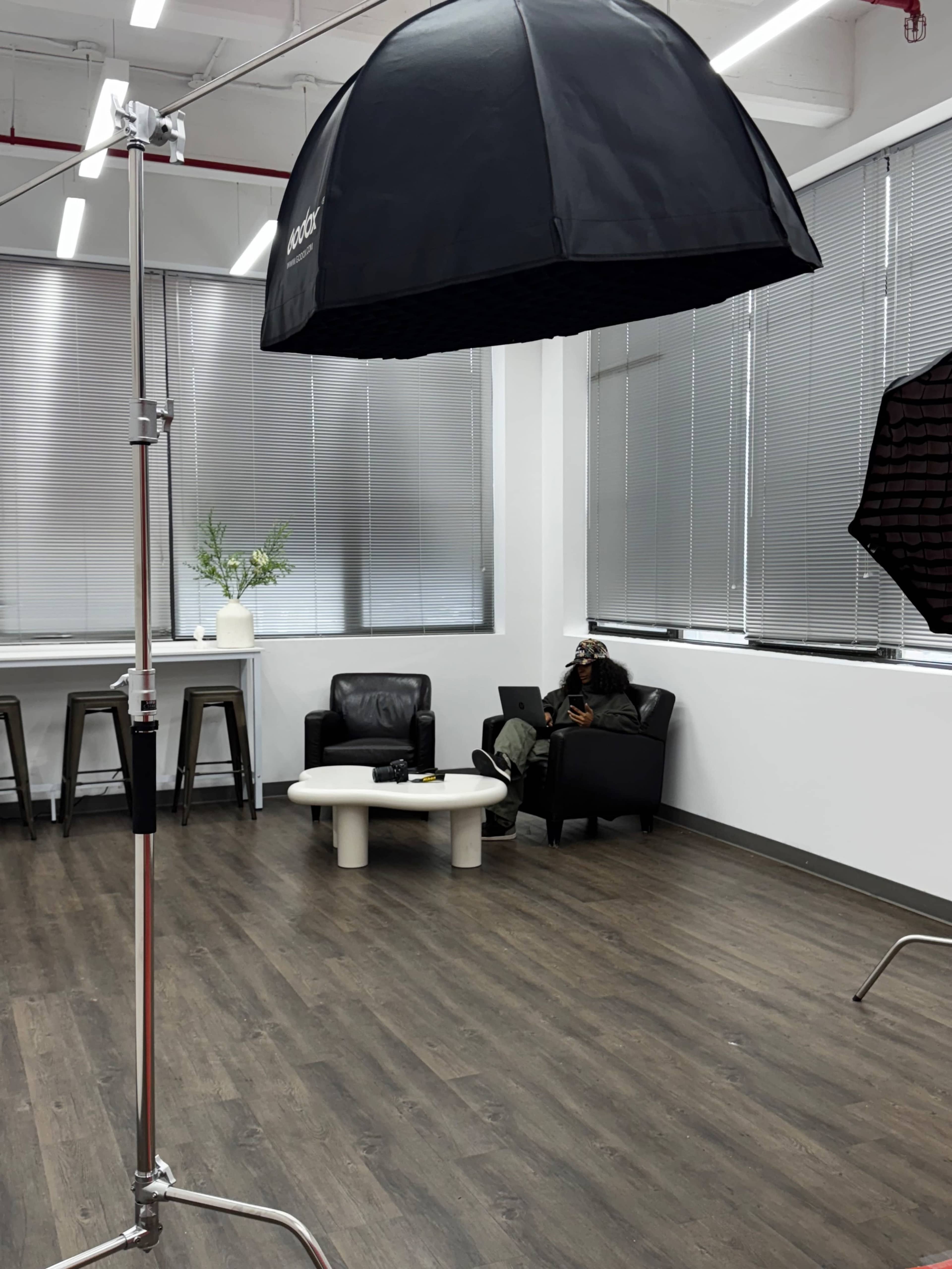 A studio space features a black umbrella light setup, a small white table, seating area with a black leather sofa, and large windows with blinds.