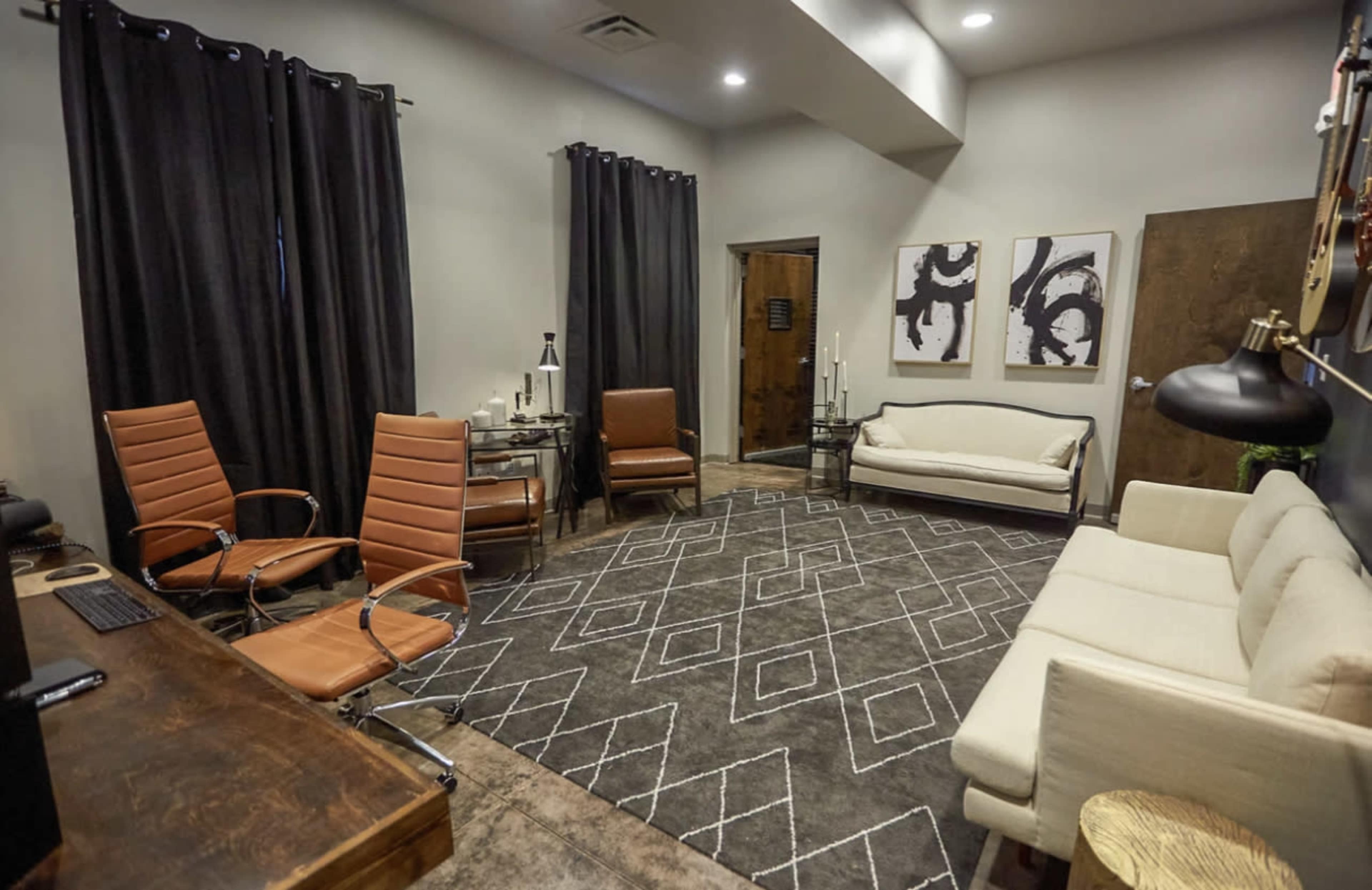A modern office space featuring a large area rug, three chairs, a sofa, a wooden desk, and two abstract artworks on the wall.