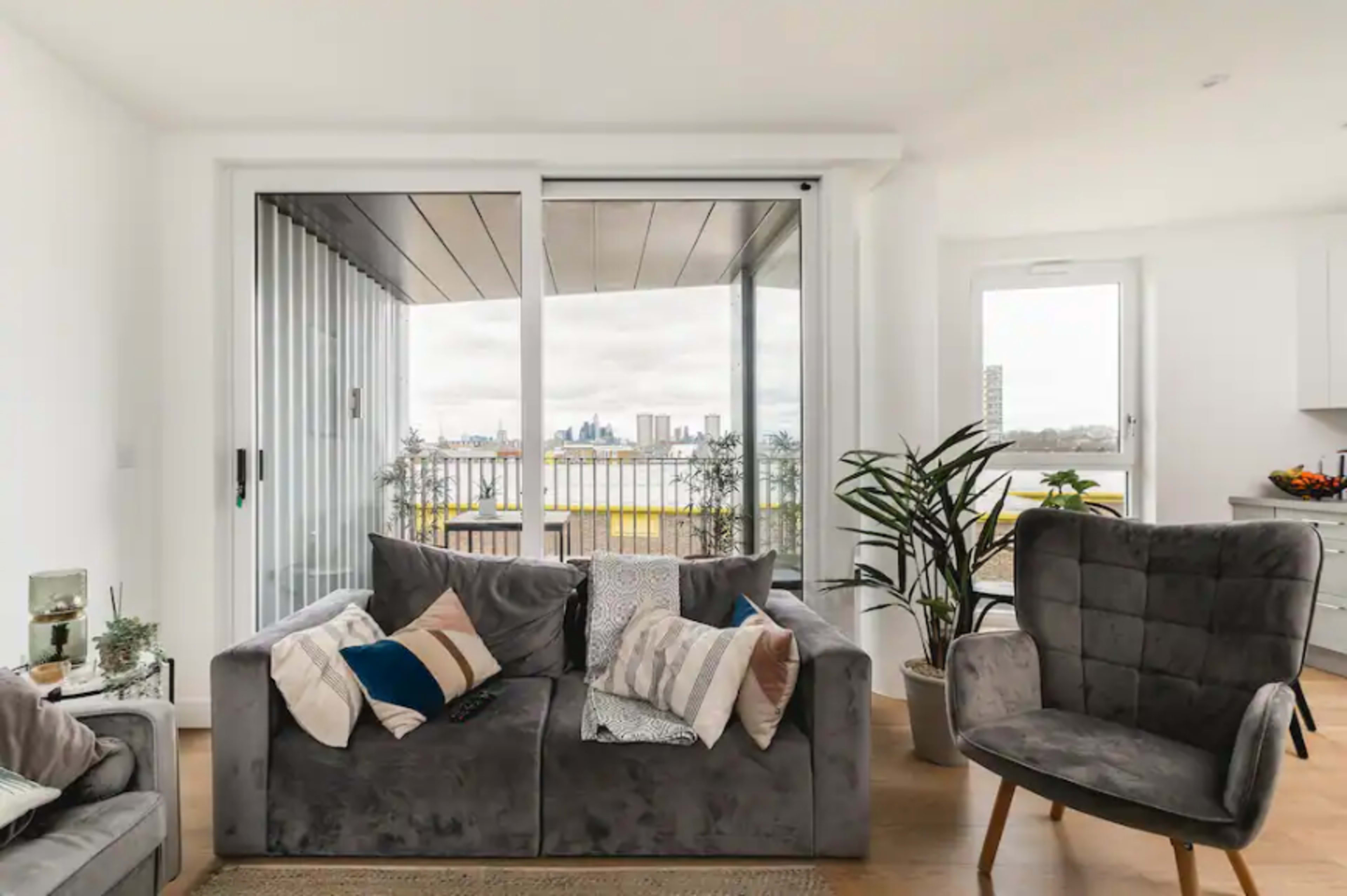 A modern living room features a gray sofa and armchair, with a view of the city visible through large glass doors leading to a balcony.