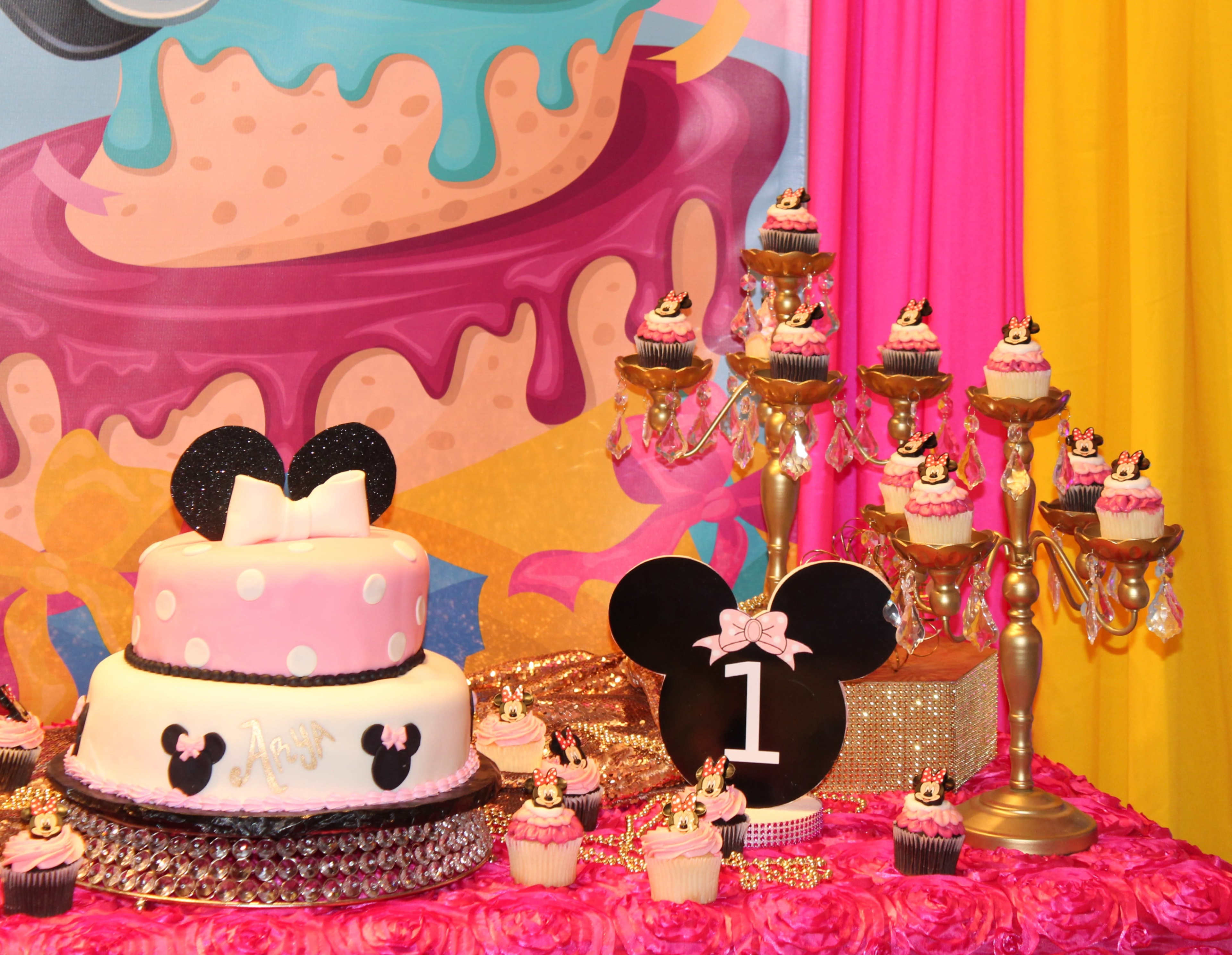 The image features a birthday dessert table decorated with a large pink and white cake, cupcakes adorned with Mickey Mouse ears, and a black number one sign.