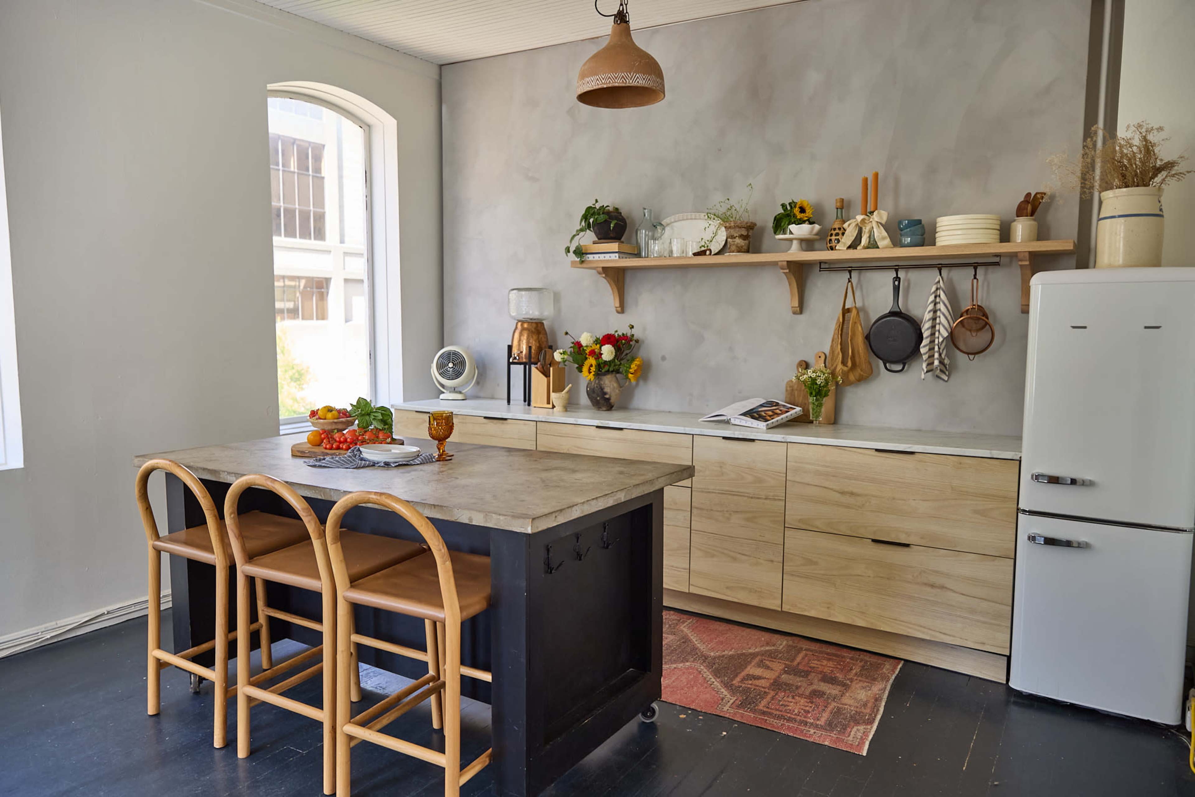 The image shows a modern kitchen featuring a concrete island with wooden bar stools, light gray walls, and open shelving displaying various kitchenware and decor.