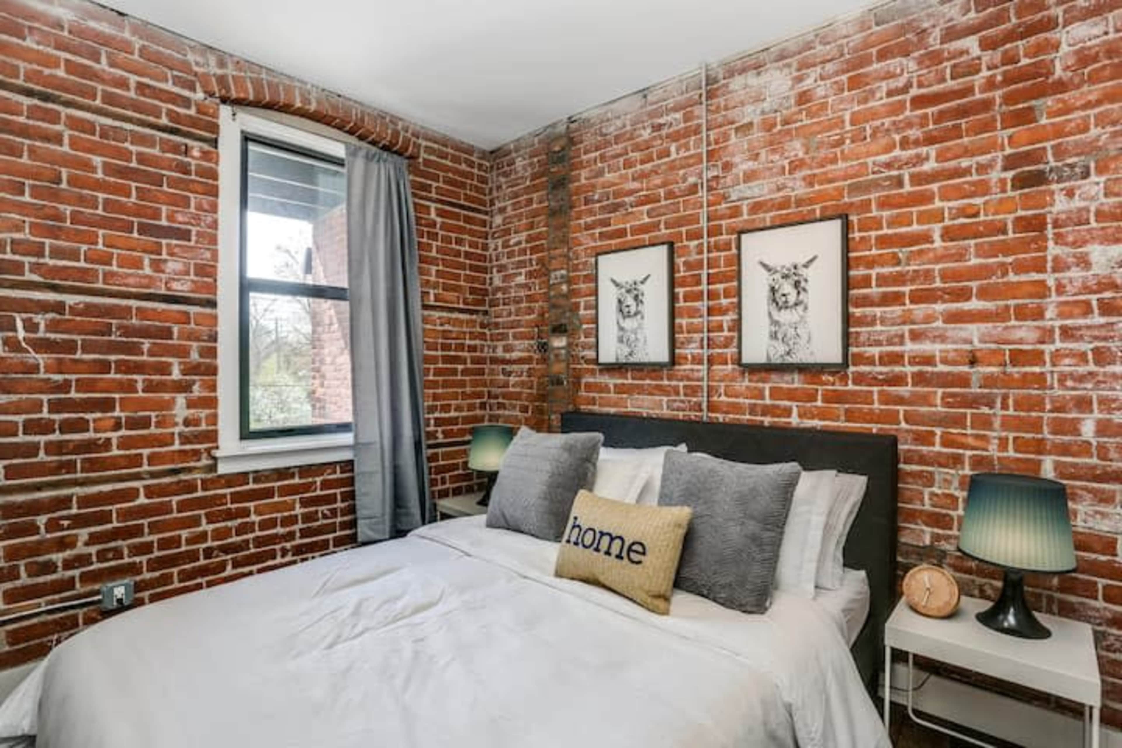 A bedroom with exposed brick walls, a bed with gray pillows and a decorative "home" pillow, and framed artwork on the walls.