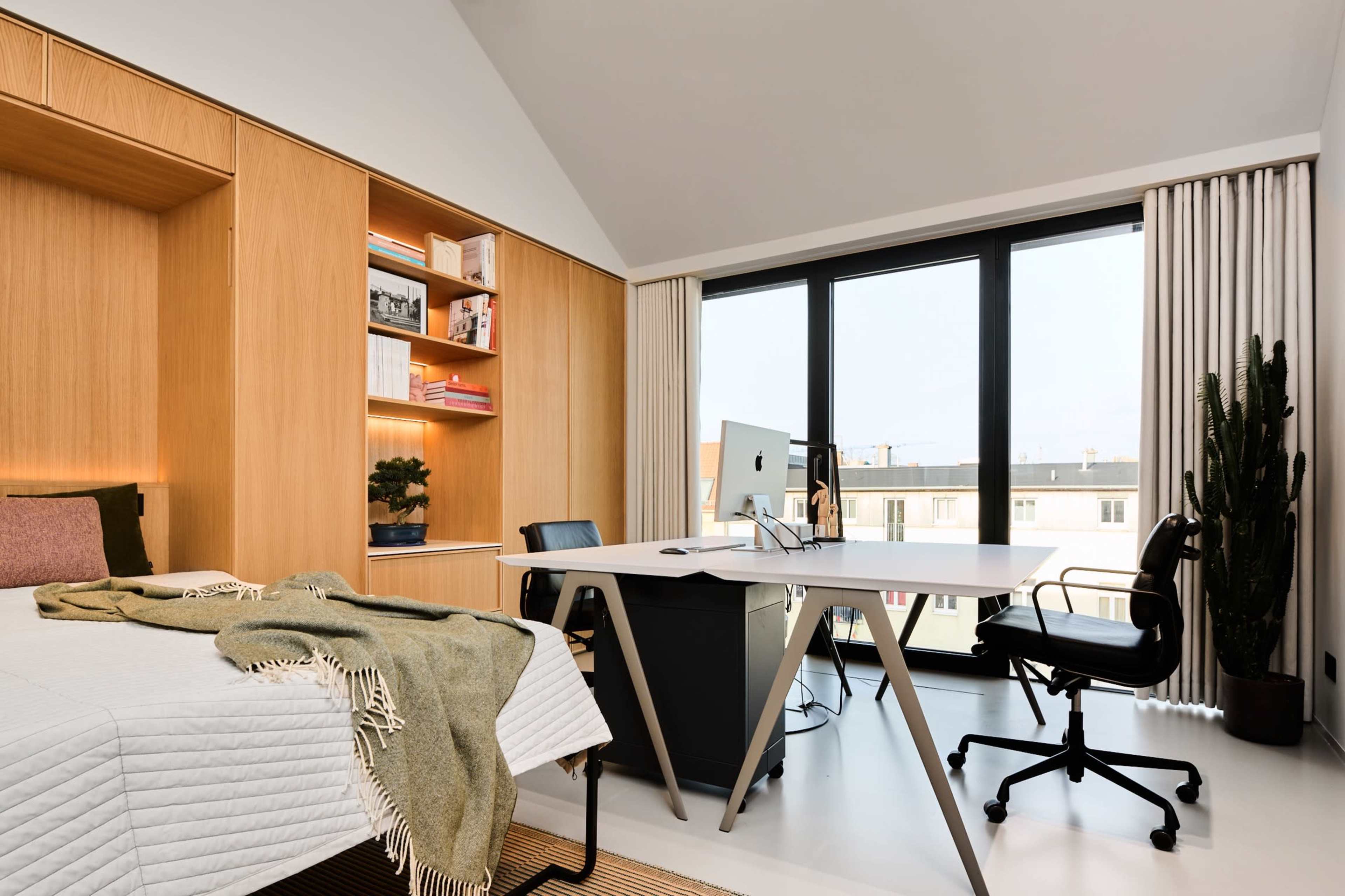 The image shows a modern bedroom with a desk and chair, featuring a large window that lets in natural light and a minimalist design with wood accents.