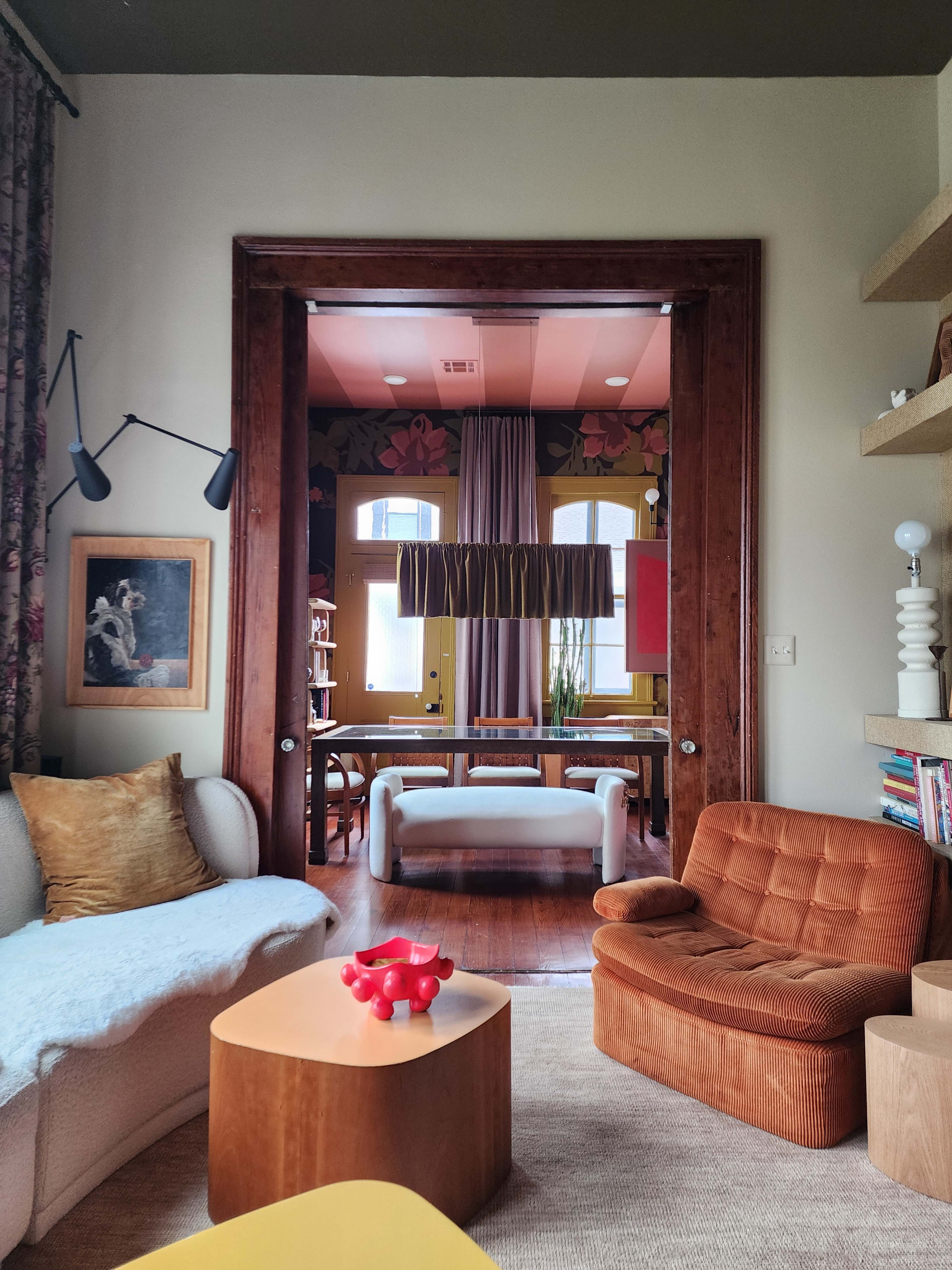 The image shows a cozy living room with a white sofa, an orange armchair, and a wooden table, leading into another room adorned with floral wallpaper and a long dining table.