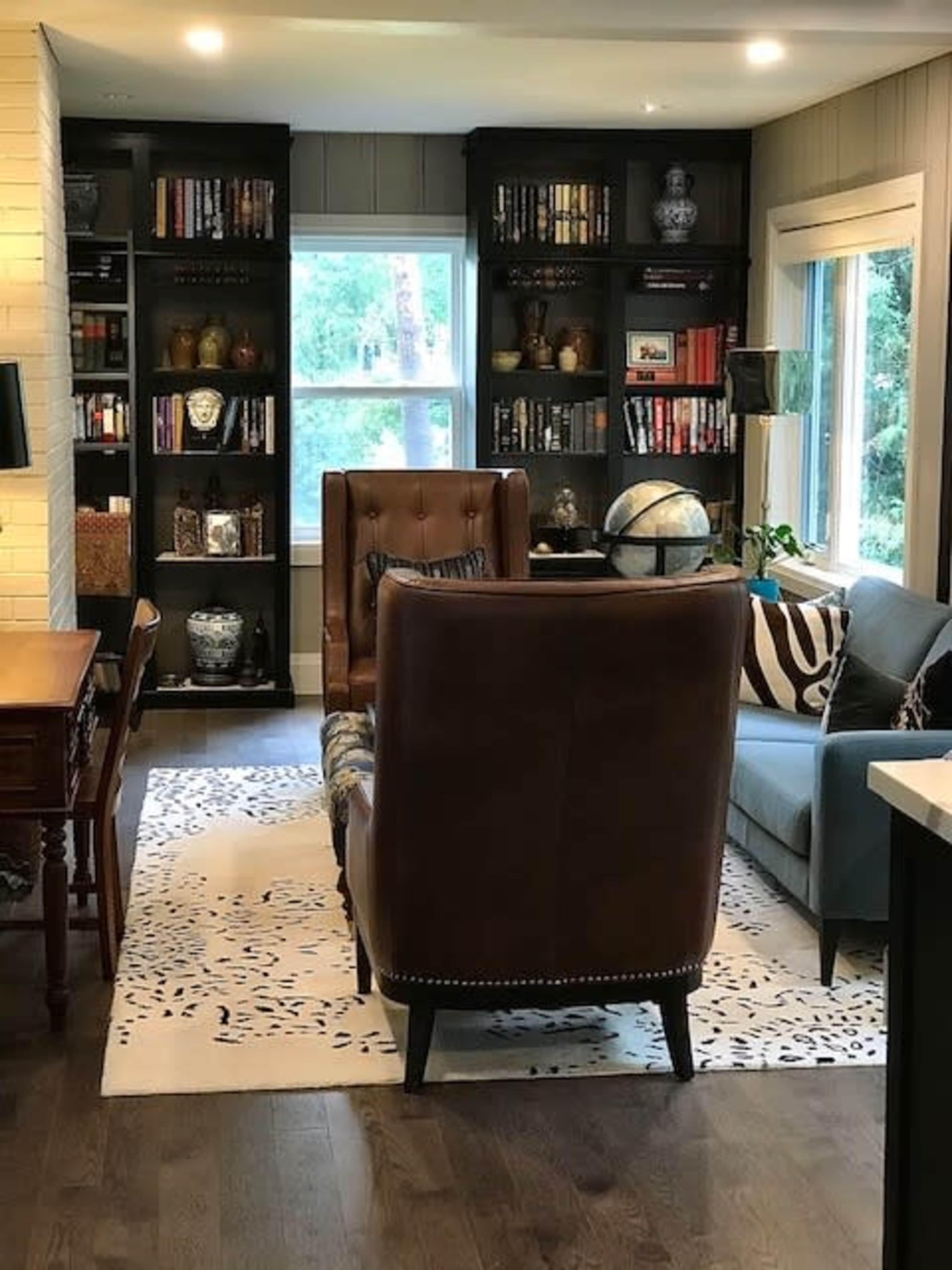 A cozy reading nook features two brown leather chairs facing a bookshelf filled with books and decorative items, with a window letting in natural light.