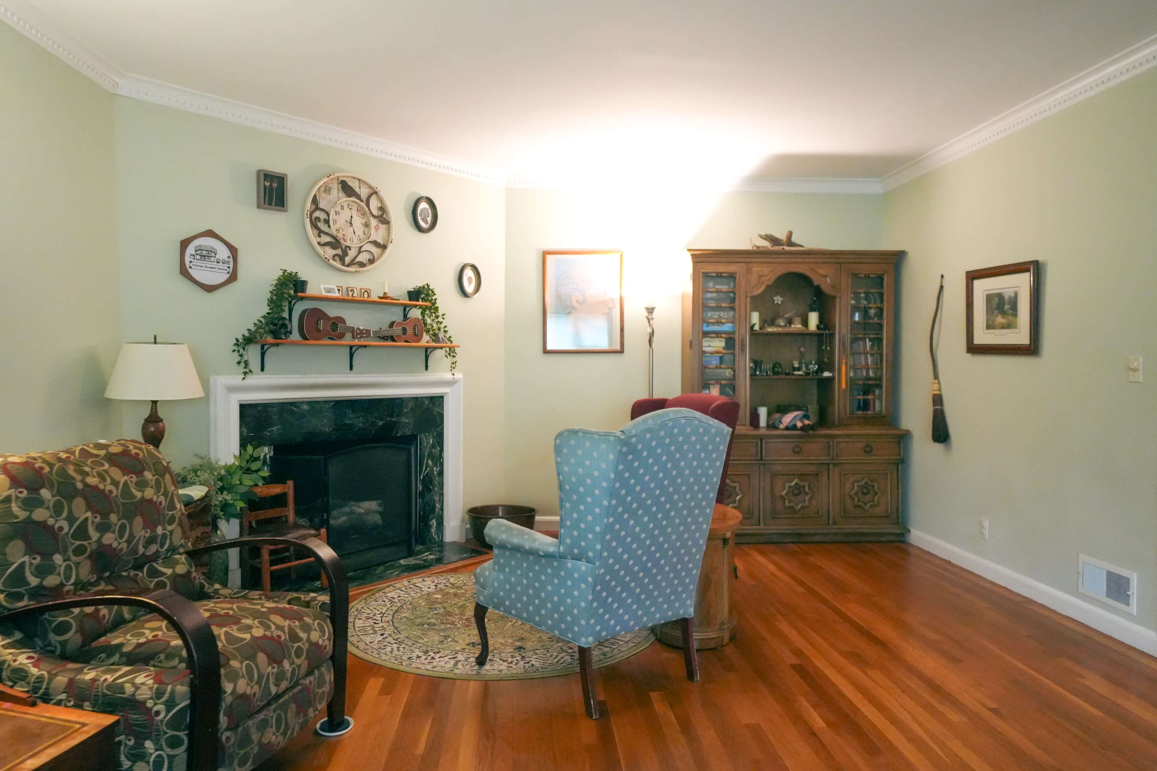 The living room features a fireplace, a wooden cabinet, and two upholstered chairs arranged around a round table on hardwood flooring.