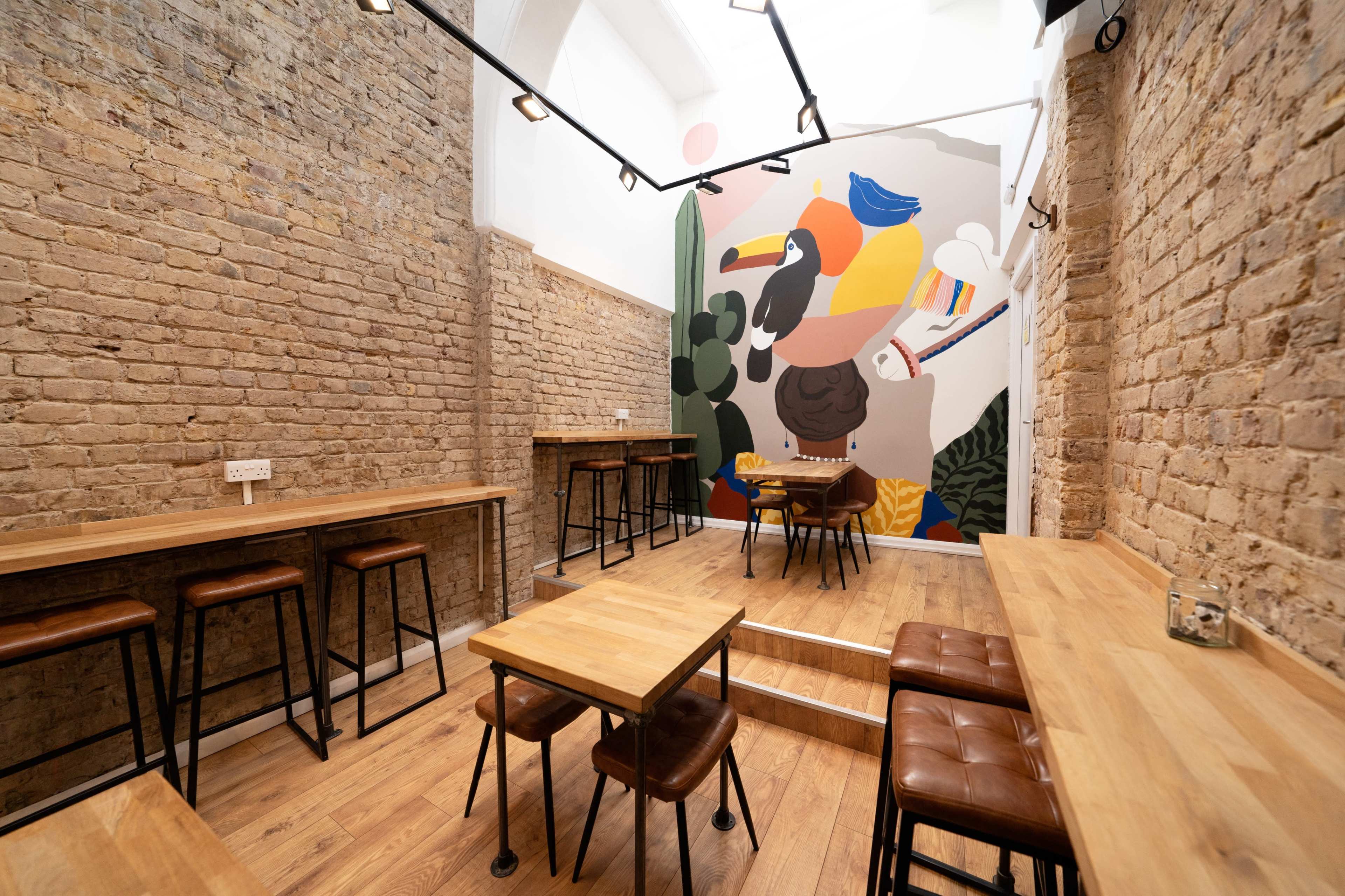 A modern cafe interior with wooden tables and stools, featuring a large colorful mural on one brick wall.