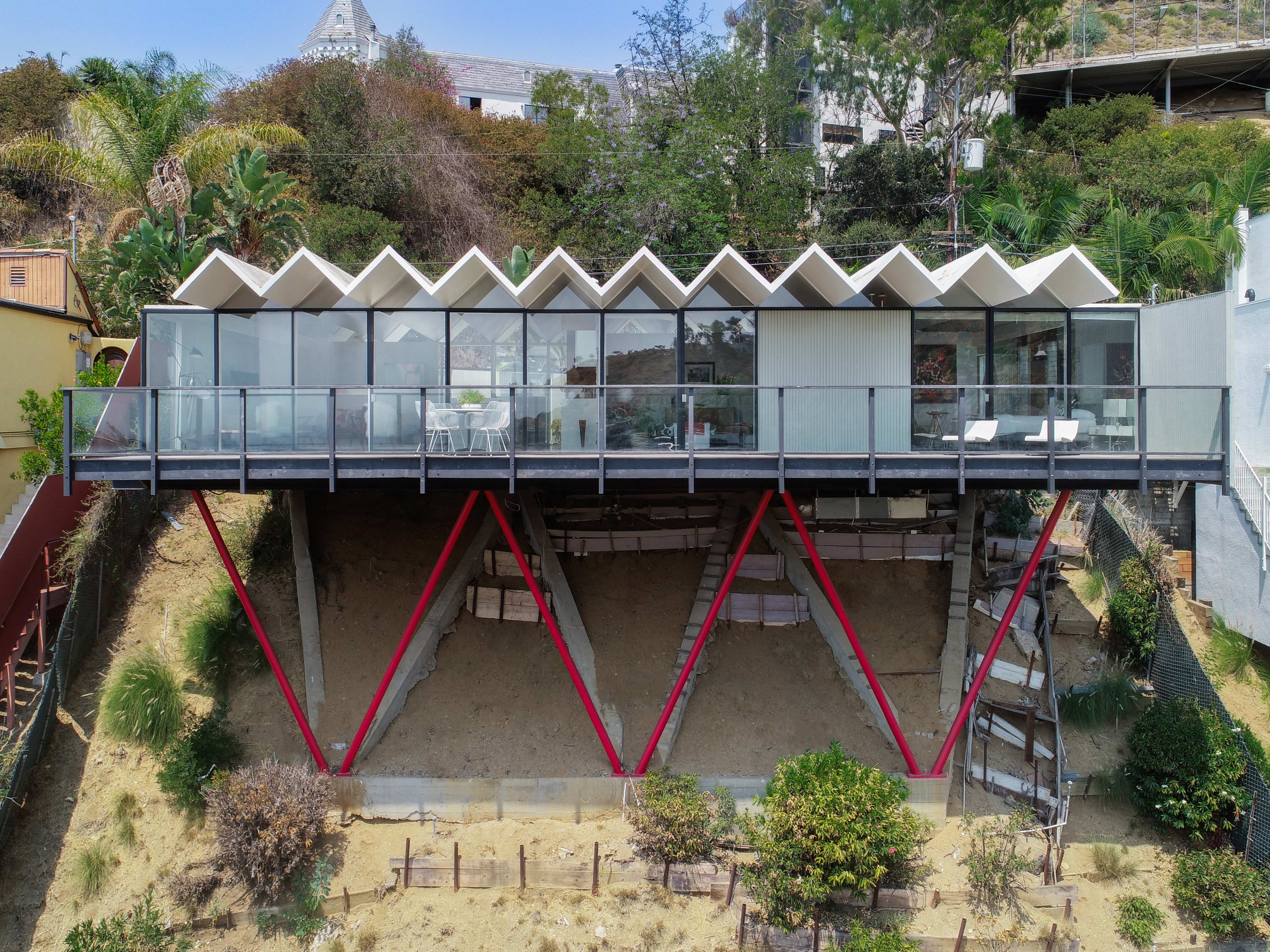 A modern house with a distinctive zigzag roof is elevated on red supports, overlooking a sandy hillside and surrounded by greenery.