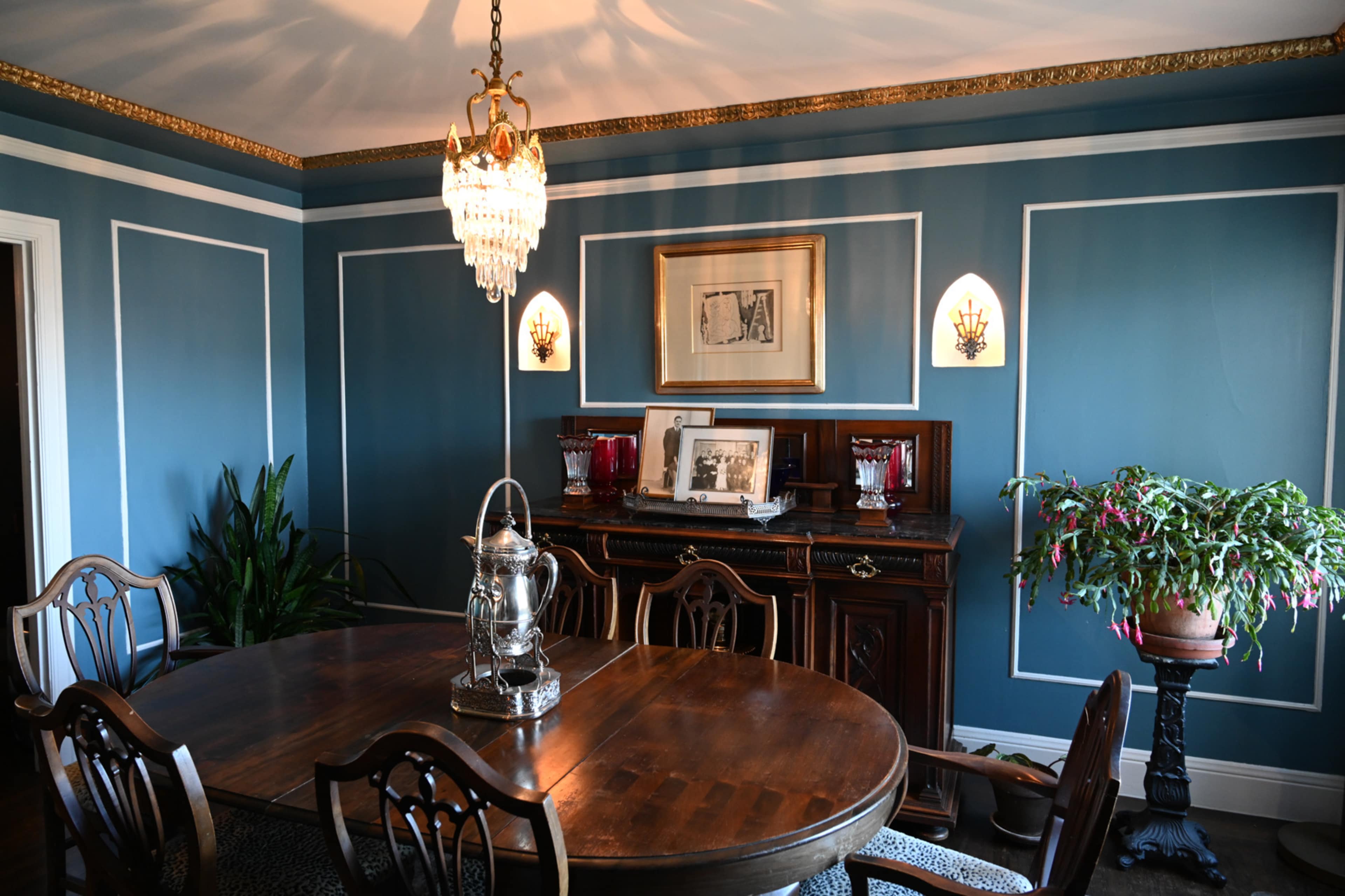A dining room with a round wooden table, a decorative chandelier, and blue walls featuring white trim, complemented by a sideboard and plant.