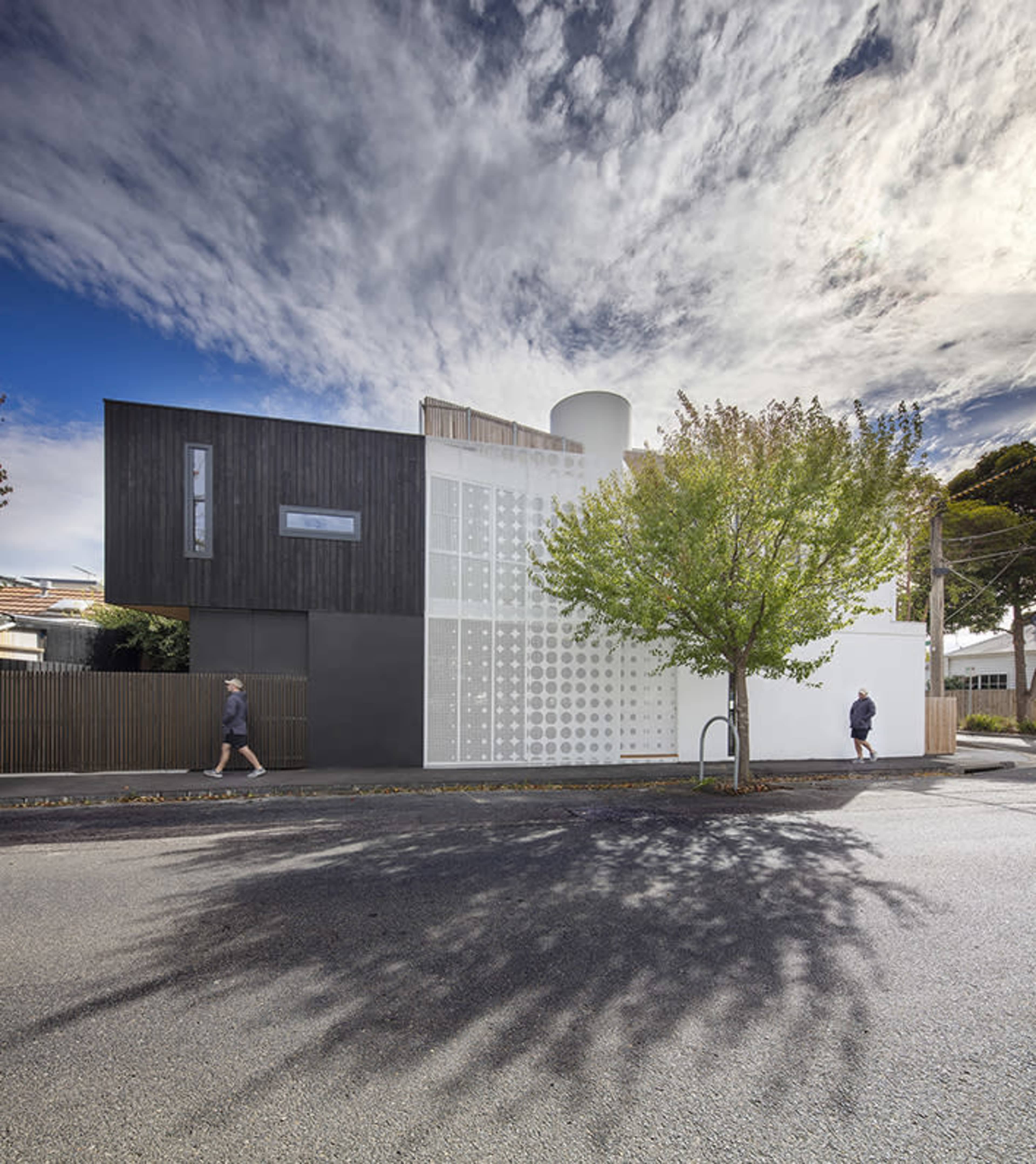 A modern two-story house with a combination of white and dark exterior materials, set against a cloudy sky and surrounded by trees and pedestrians on the street.