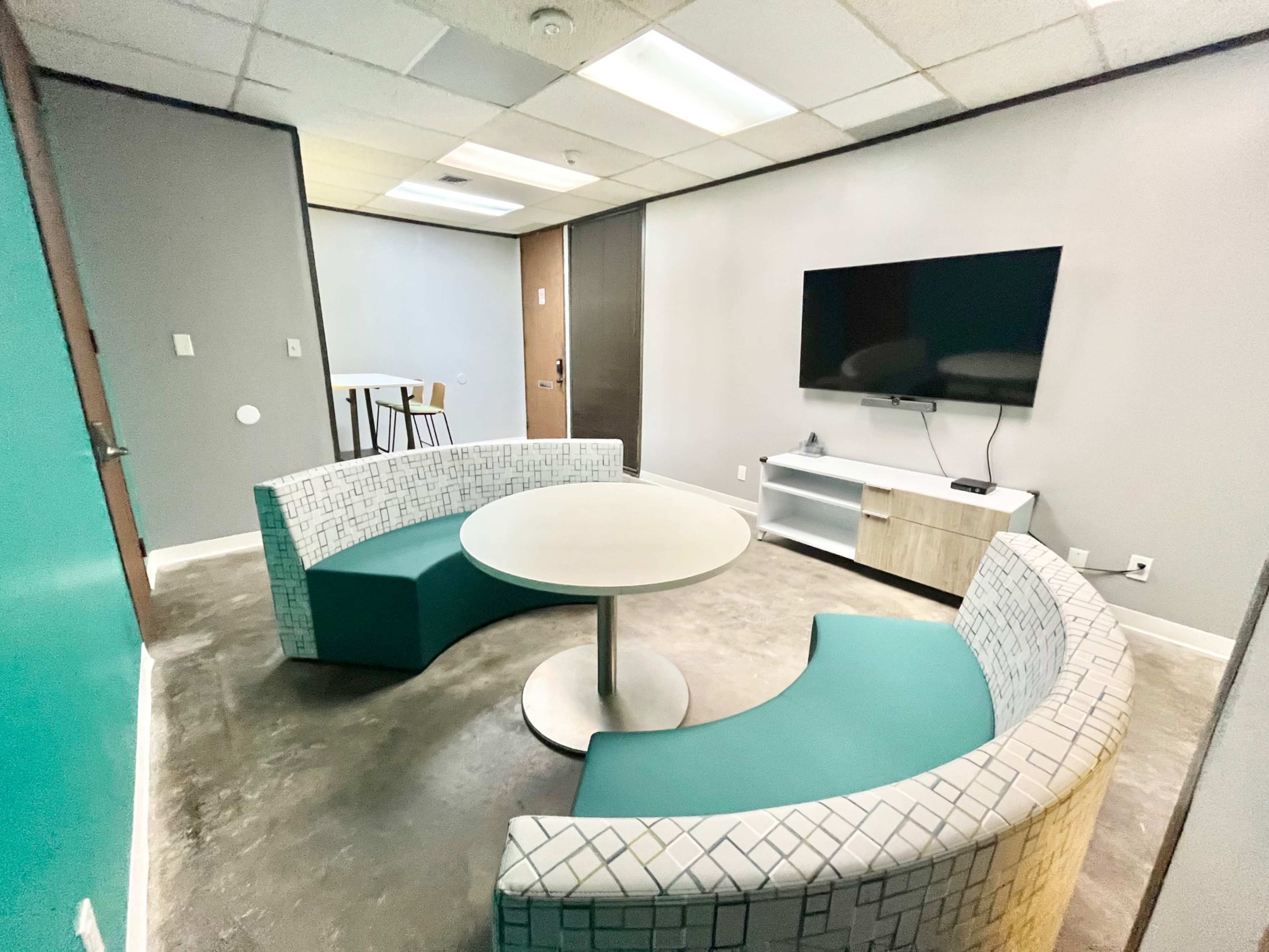 The image shows a small, modern lounge room with a circular table and curved seating, along with a television mounted on the wall.