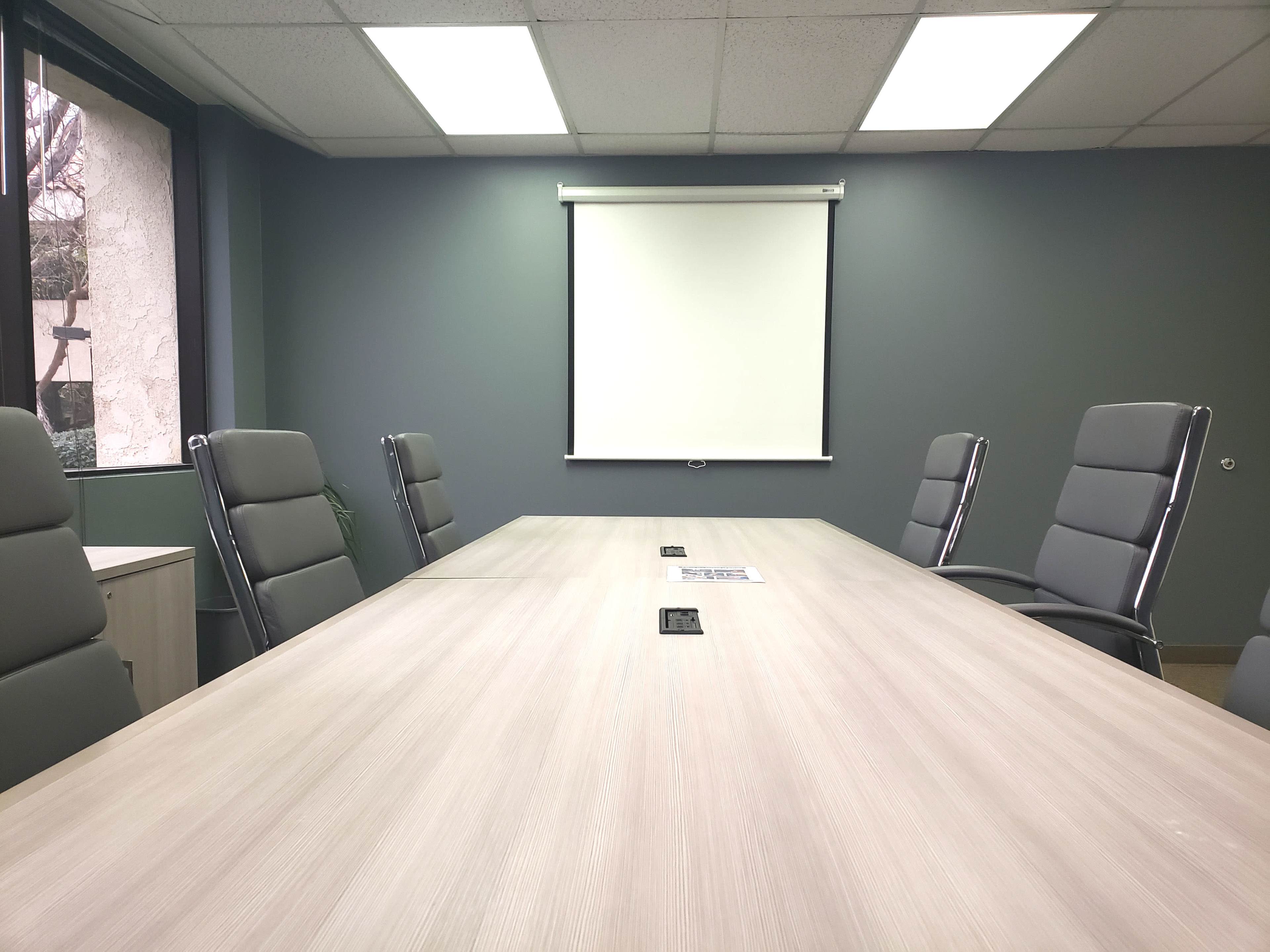A conference room with a wooden table, six gray office chairs, and a white projection screen on a green wall.
