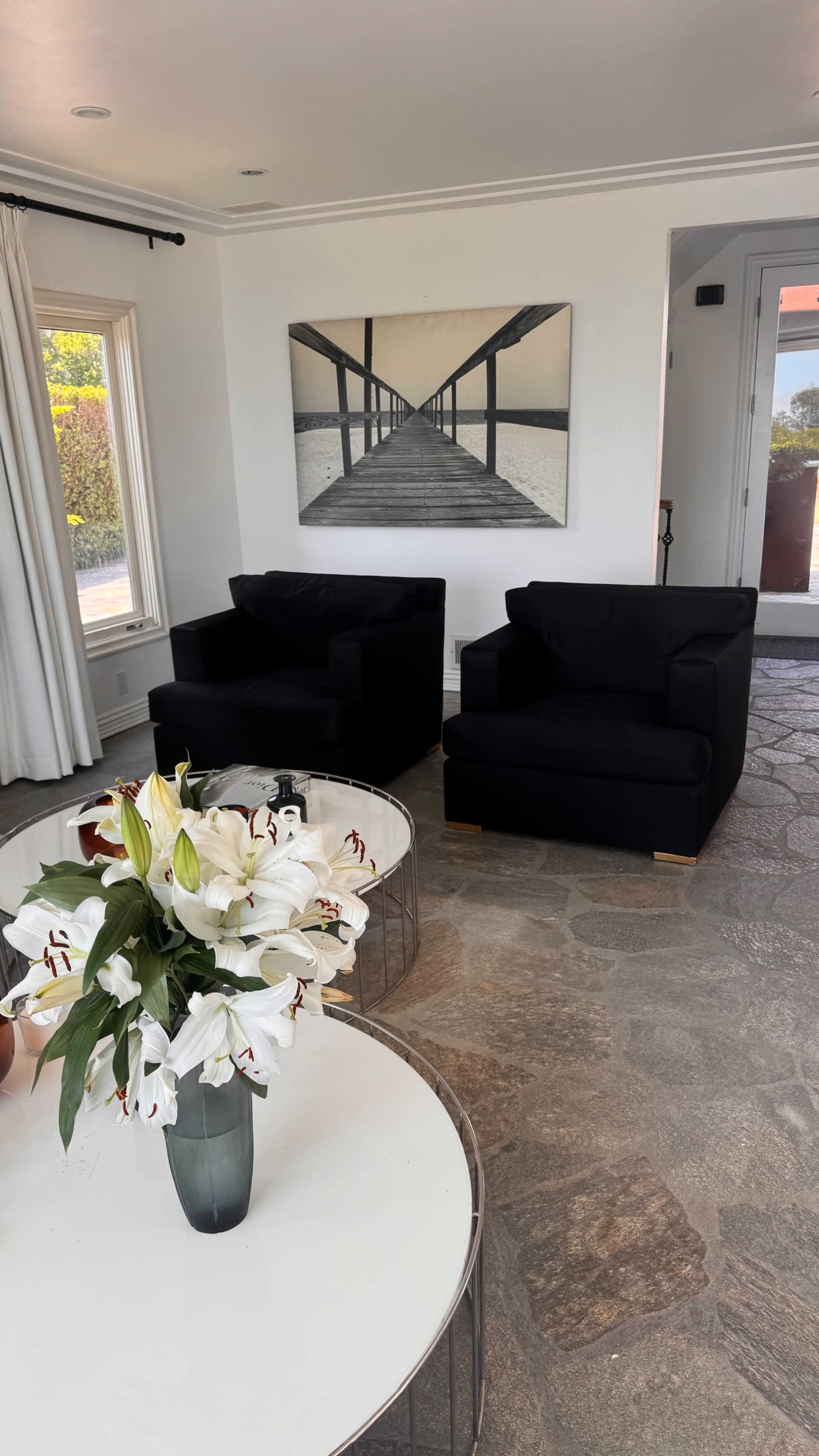 The image shows a modern living room featuring two black armchairs, a round coffee table with a vase of white lilies, and a large photograph of a pier on the wall.