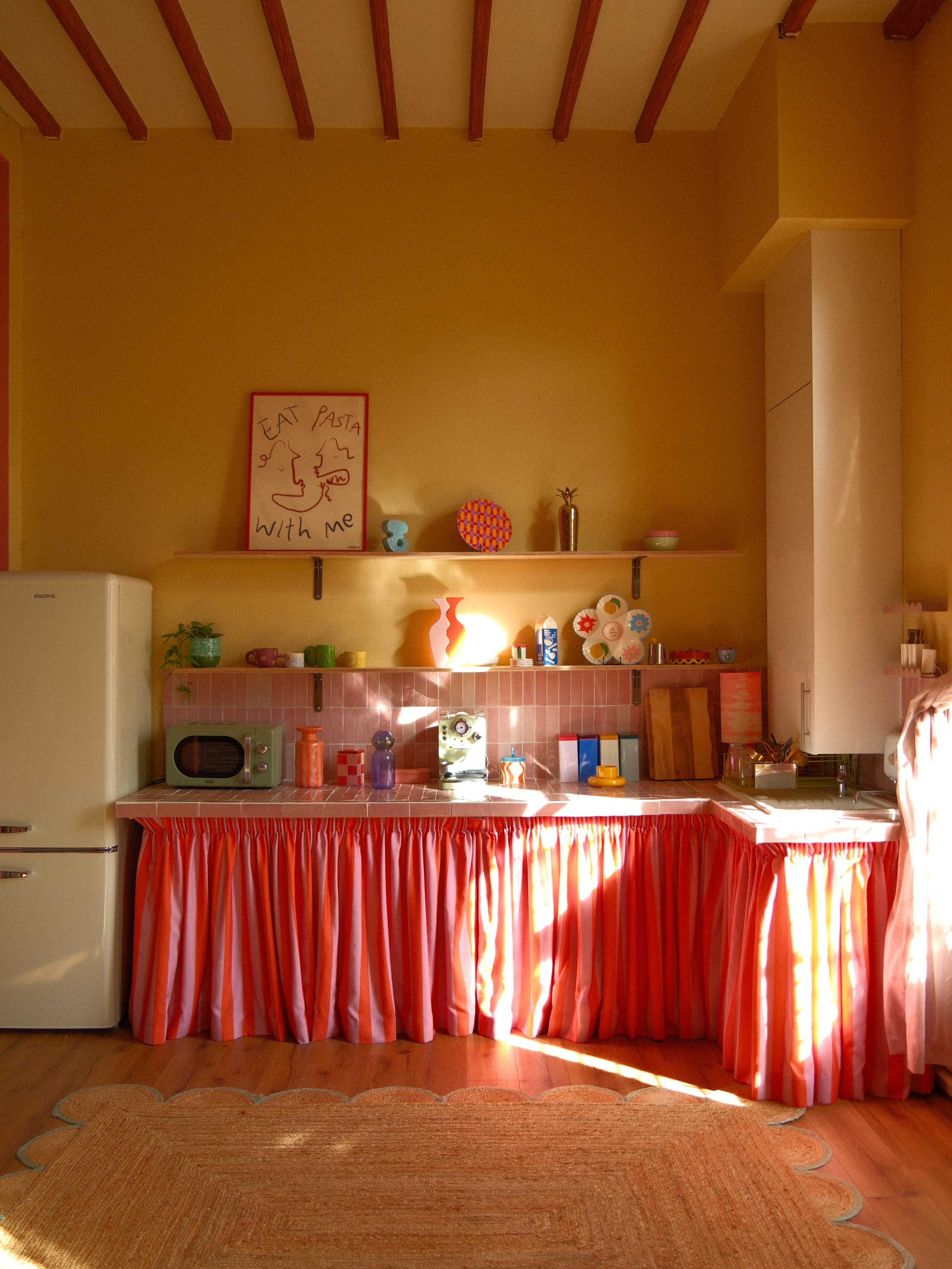 The kitchen features a colorful design with a retro refrigerator, a pink ruffled skirt around the countertop, and various kitchen items neatly arranged on shelves.
