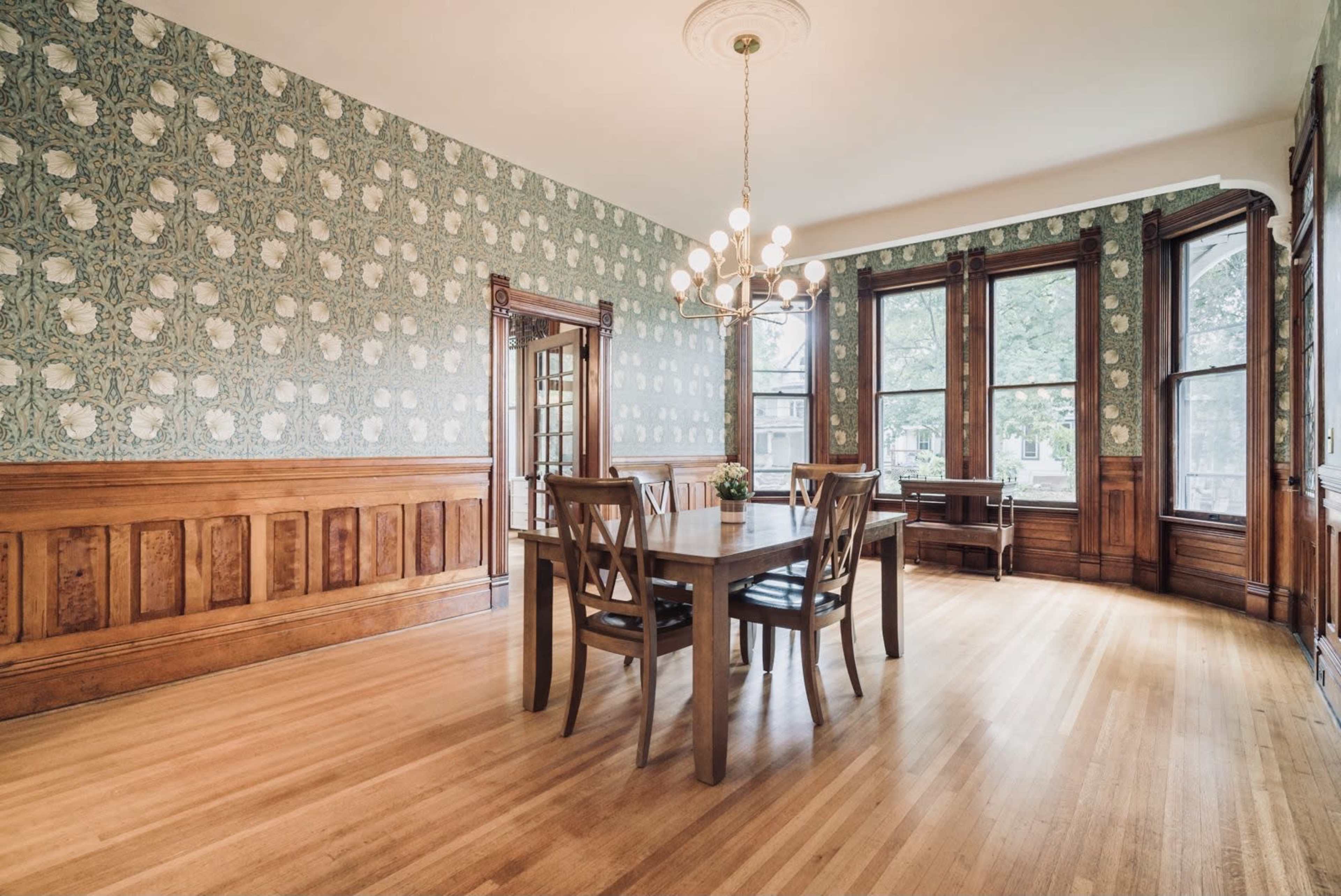 The dining room features a wooden dining table surrounded by chairs and is adorned with patterned wallpaper and large windows.