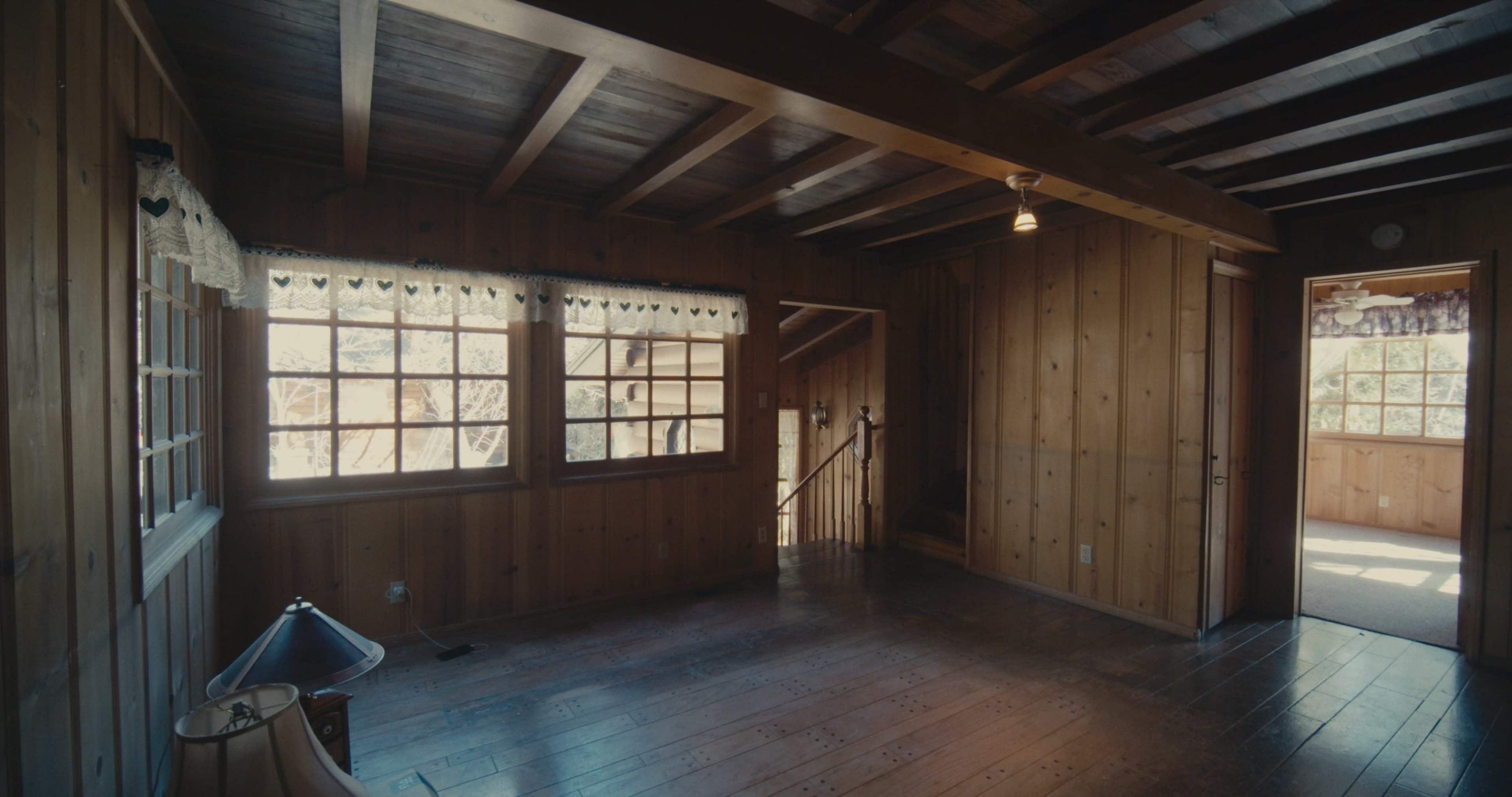 The image shows a wooden interior room with large windows, a staircase, and a lamp in the corner.