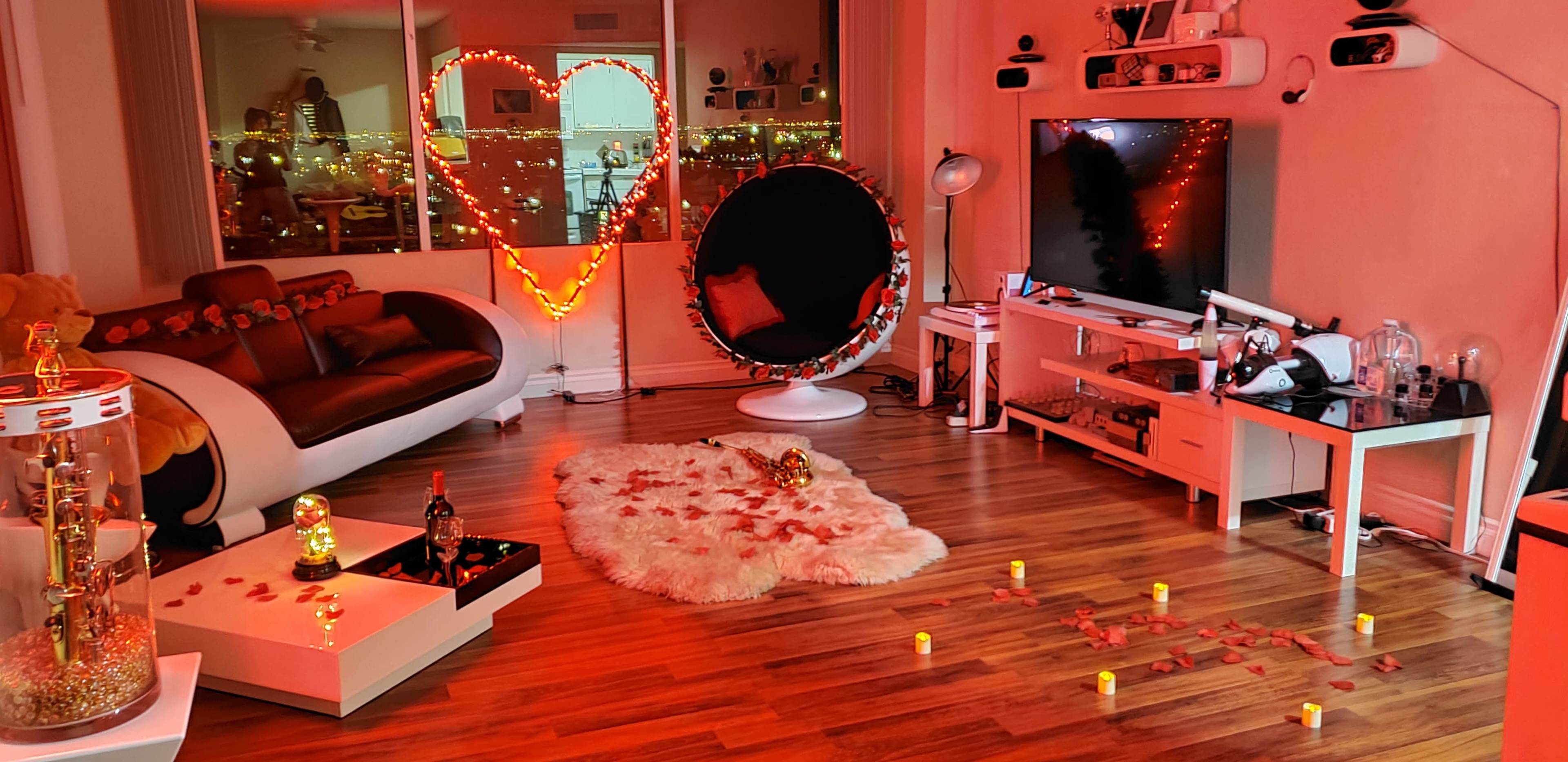The living room features a heart-shaped arrangement of lights, a cozy chair, and decorative candles on the wooden floor, surrounded by rose petals.