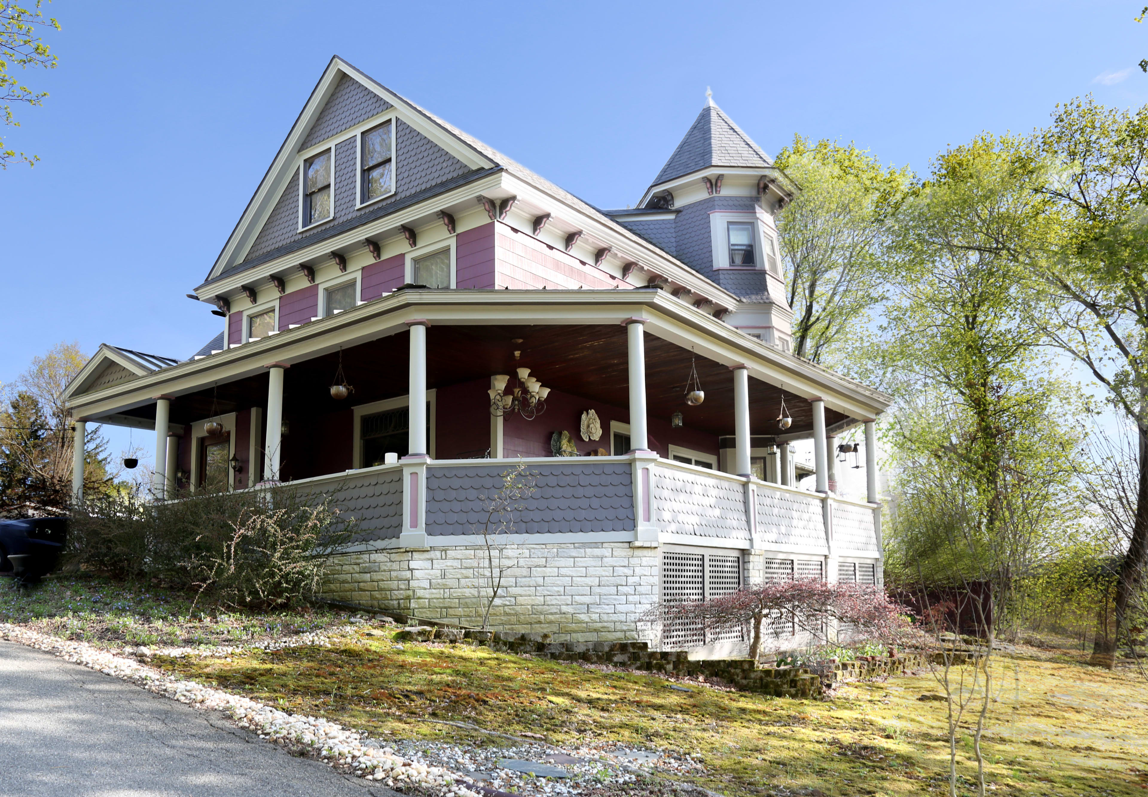 The image shows a large Victorian-style house with a tower, intricate details, and a wraparound porch surrounded by greenery.