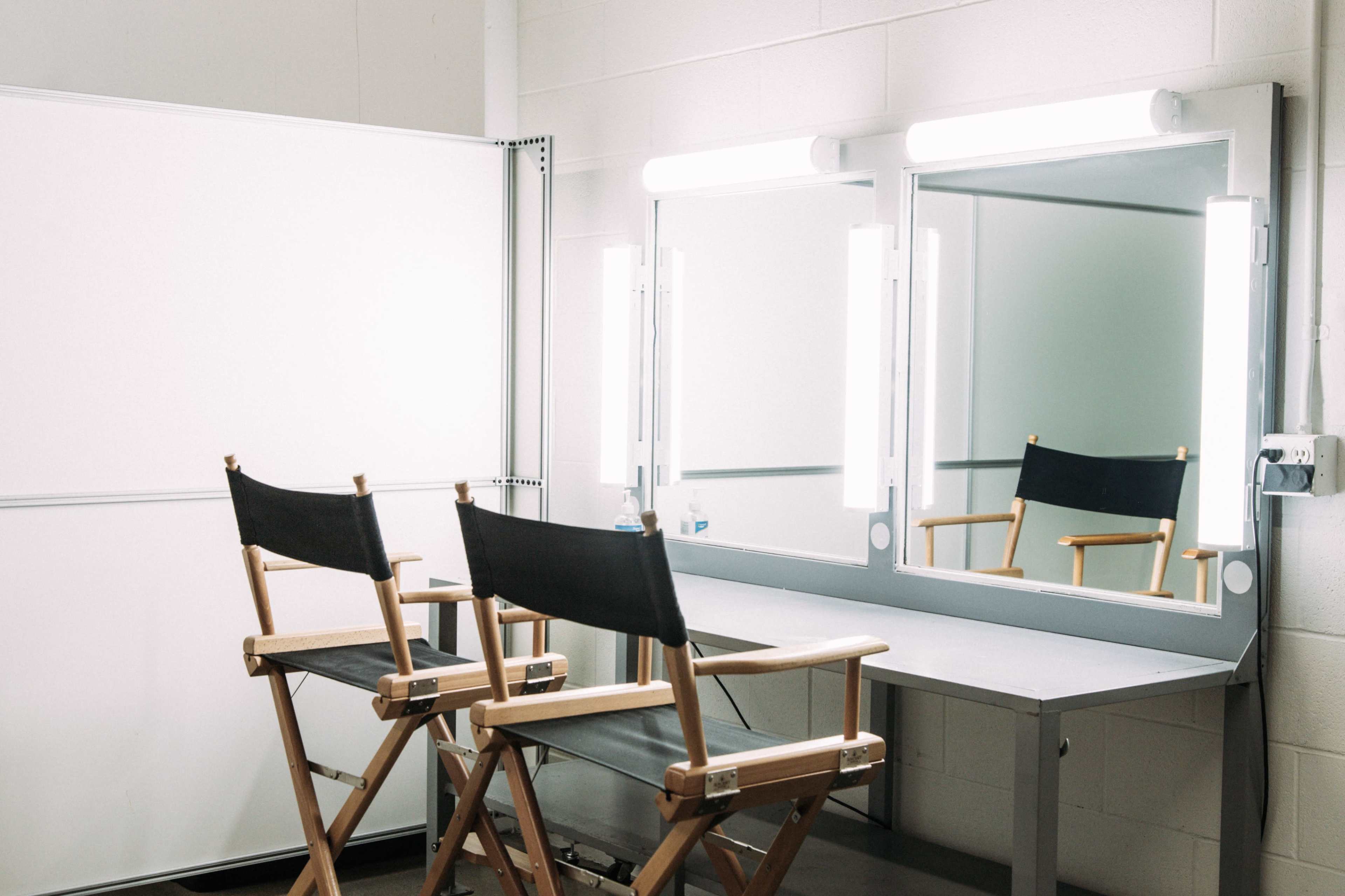 A makeup studio featuring two director's chairs in front of a well-lit mirror and a simple table.