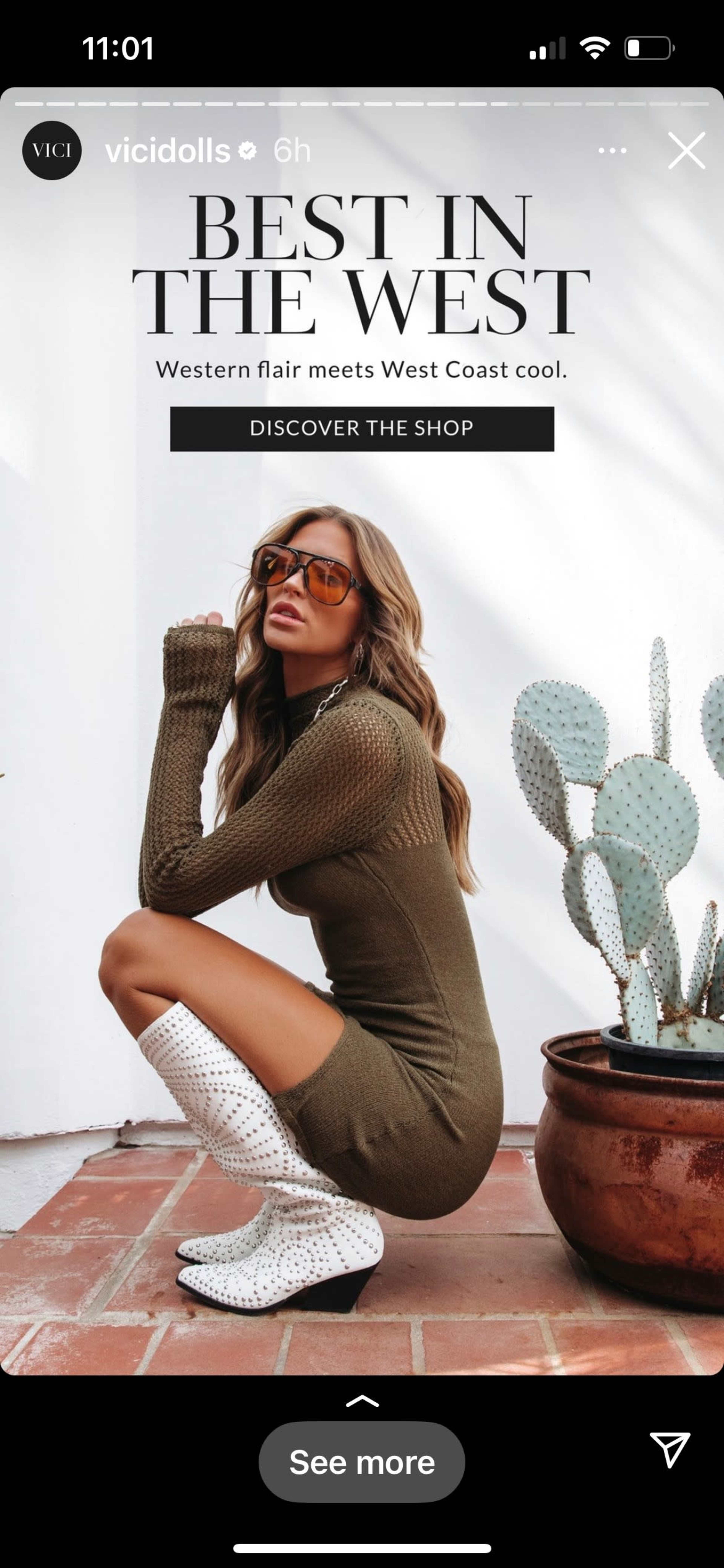 A woman in a form-fitting olive dress poses beside a potted cactus while wearing knee-high, white speckled boots and oversized sunglasses.