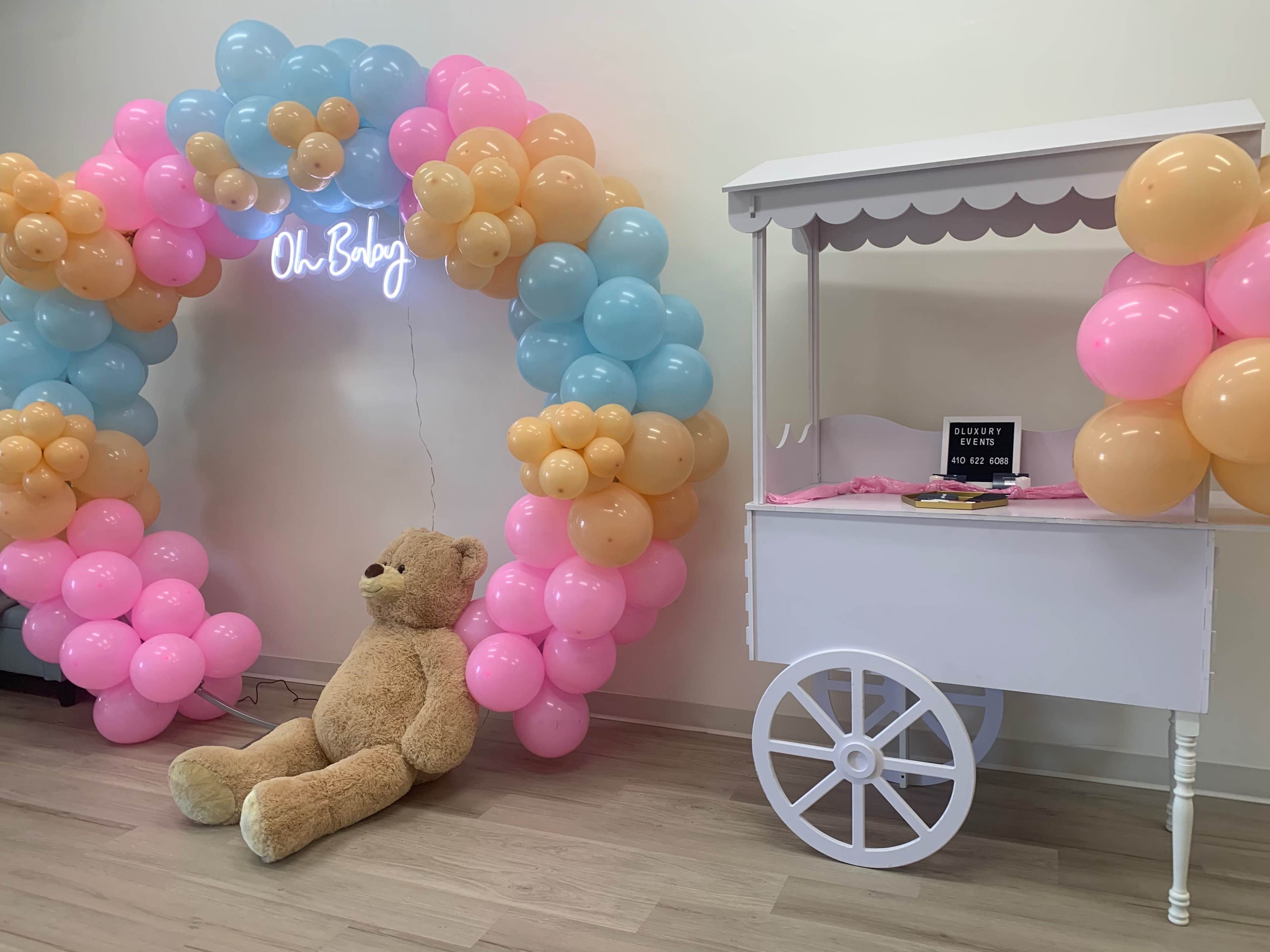 A colorful balloon arch of pink, blue, and yellow surrounds a large teddy bear sitting next to a white snack cart.