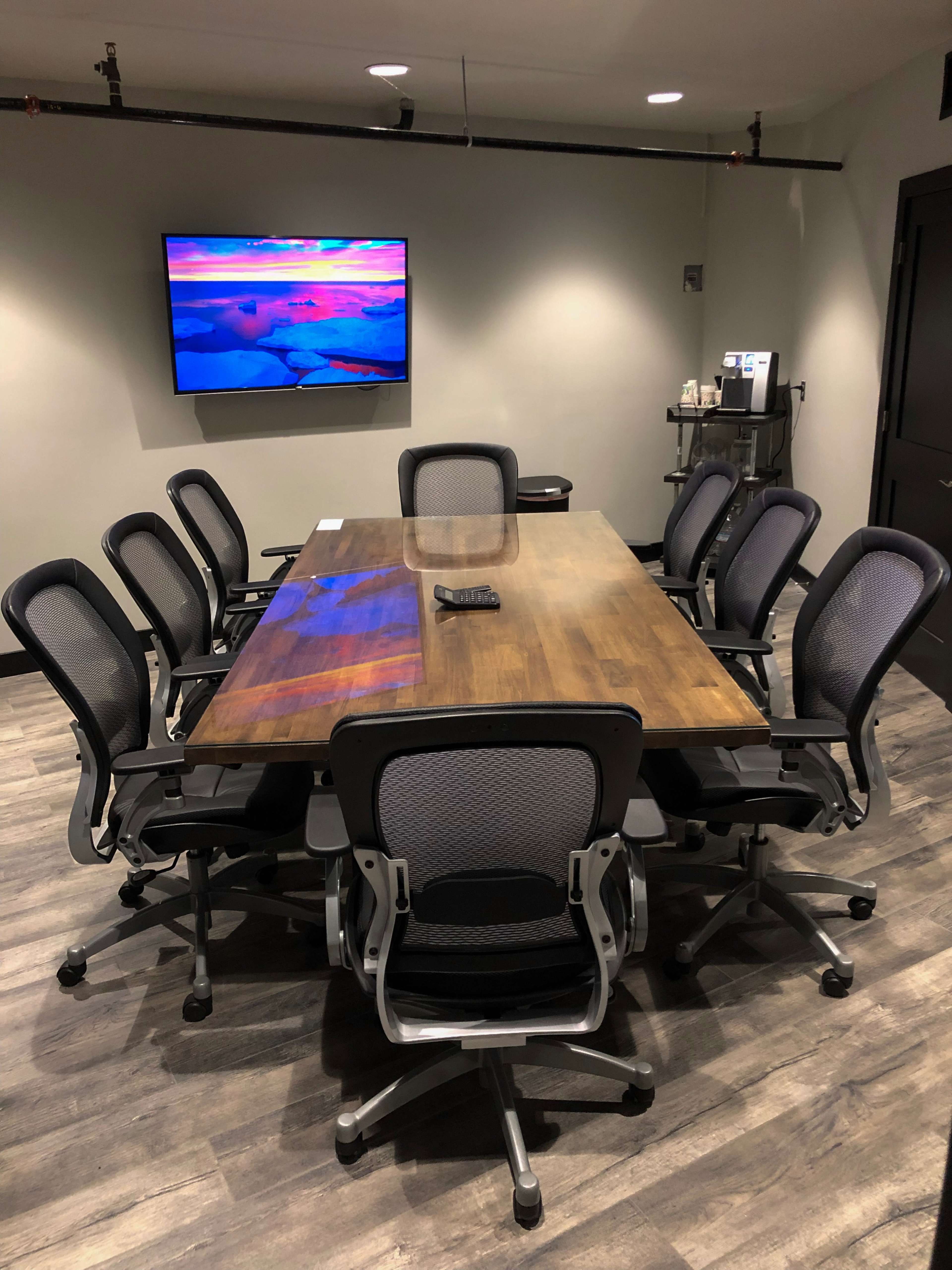 A large wooden conference table surrounded by eight office chairs faces a wall-mounted television in a meeting room.