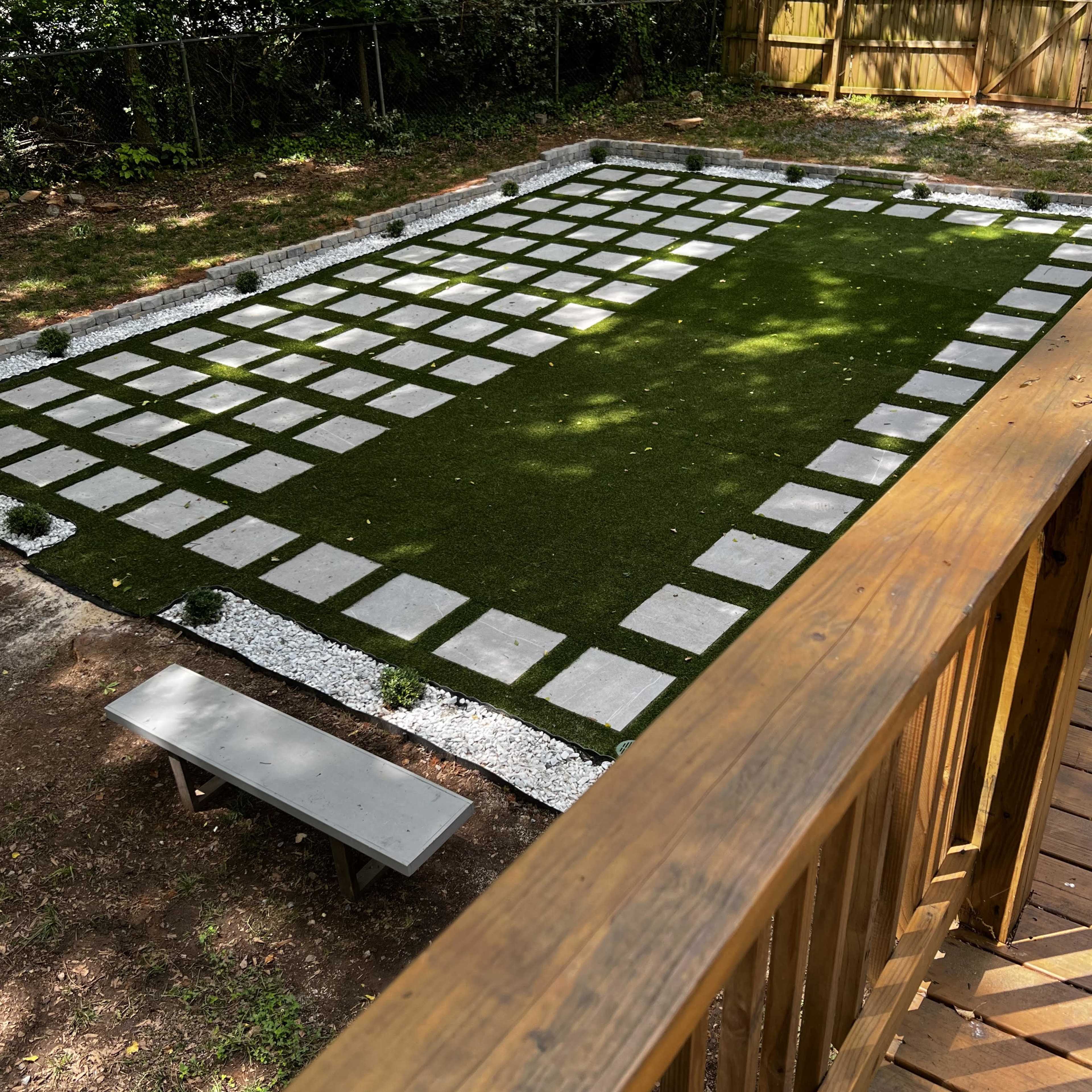 A landscaped yard featuring a grid of stone pavers on artificial grass, surrounded by white gravel and small plants.