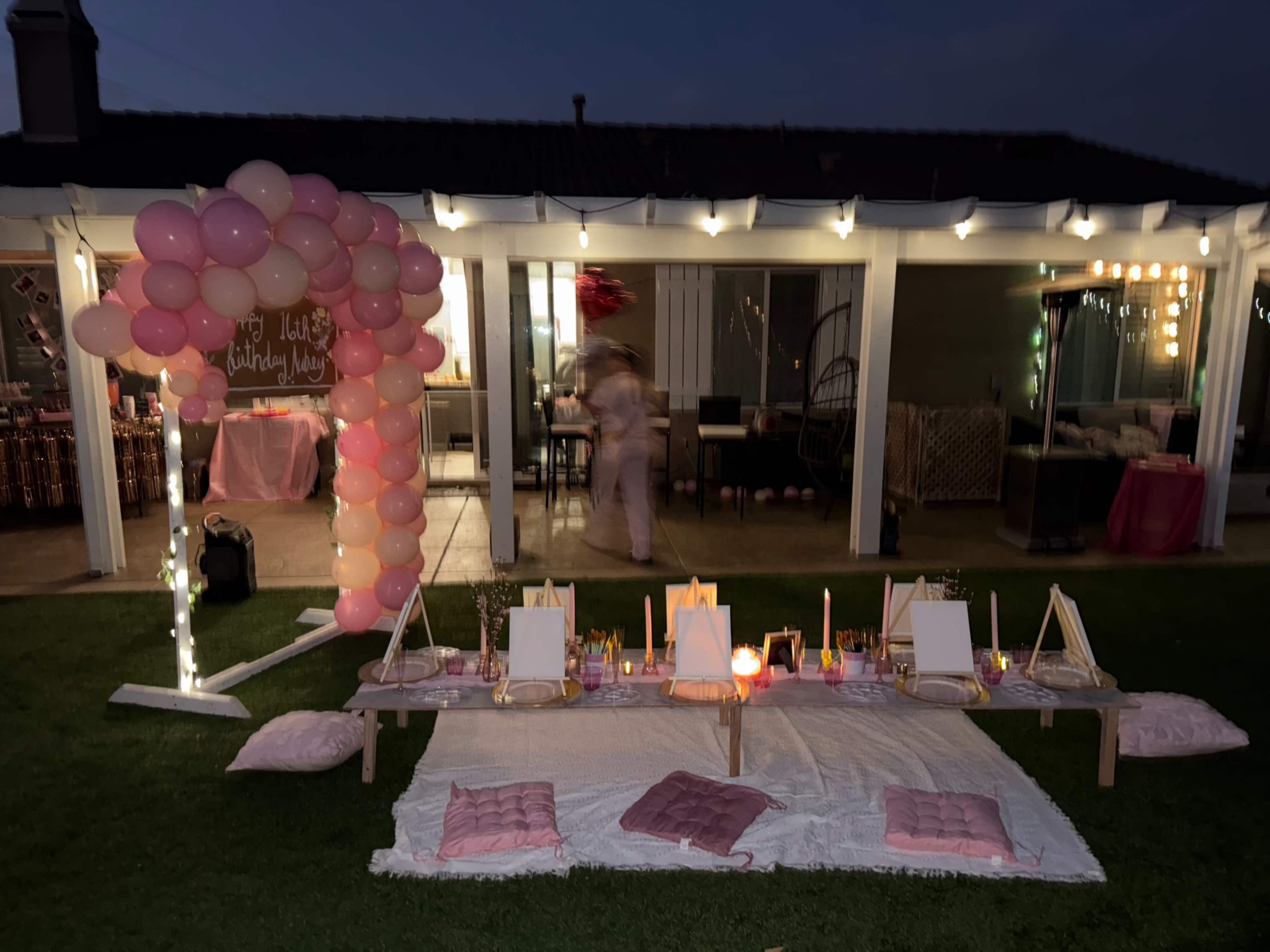 The image shows an outdoor setup for a celebration featuring a pink balloon arch, a low table with art supplies, and decorative lighting in the background.