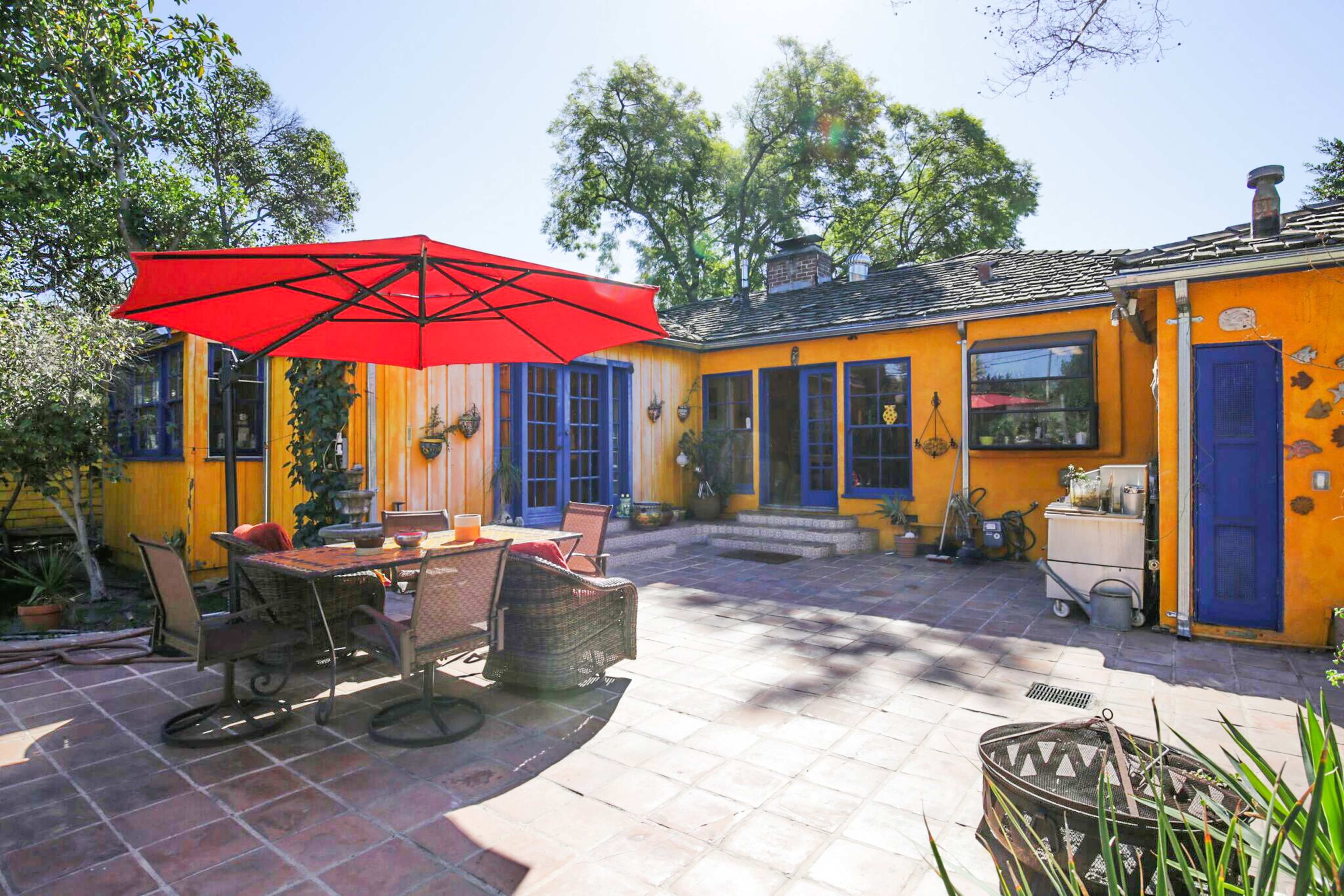 A bright yellow house with blue doors and a red patio umbrella overlooks a tiled outdoor seating area surrounded by trees.