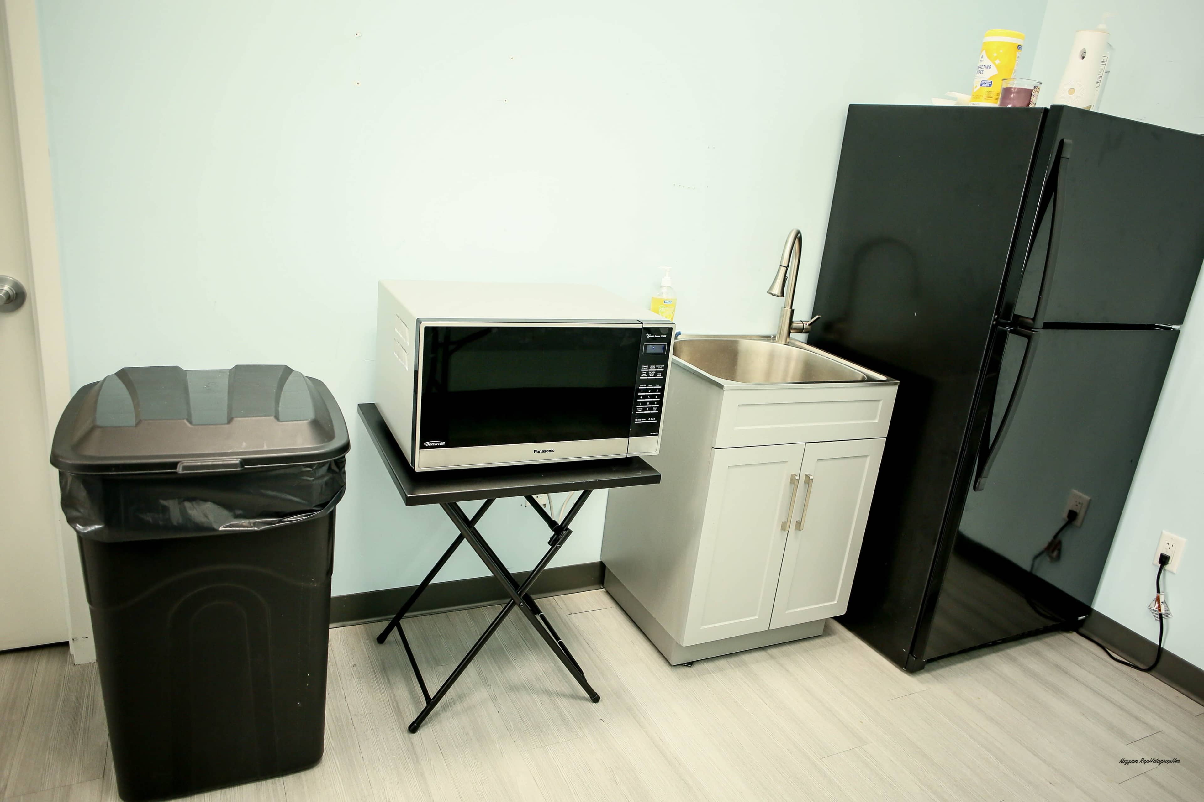 The image shows a small kitchenette featuring a black trash can, a microwave on a folding table, a stainless steel sink with a cabinet, and a black refrigerator against a light blue wall.