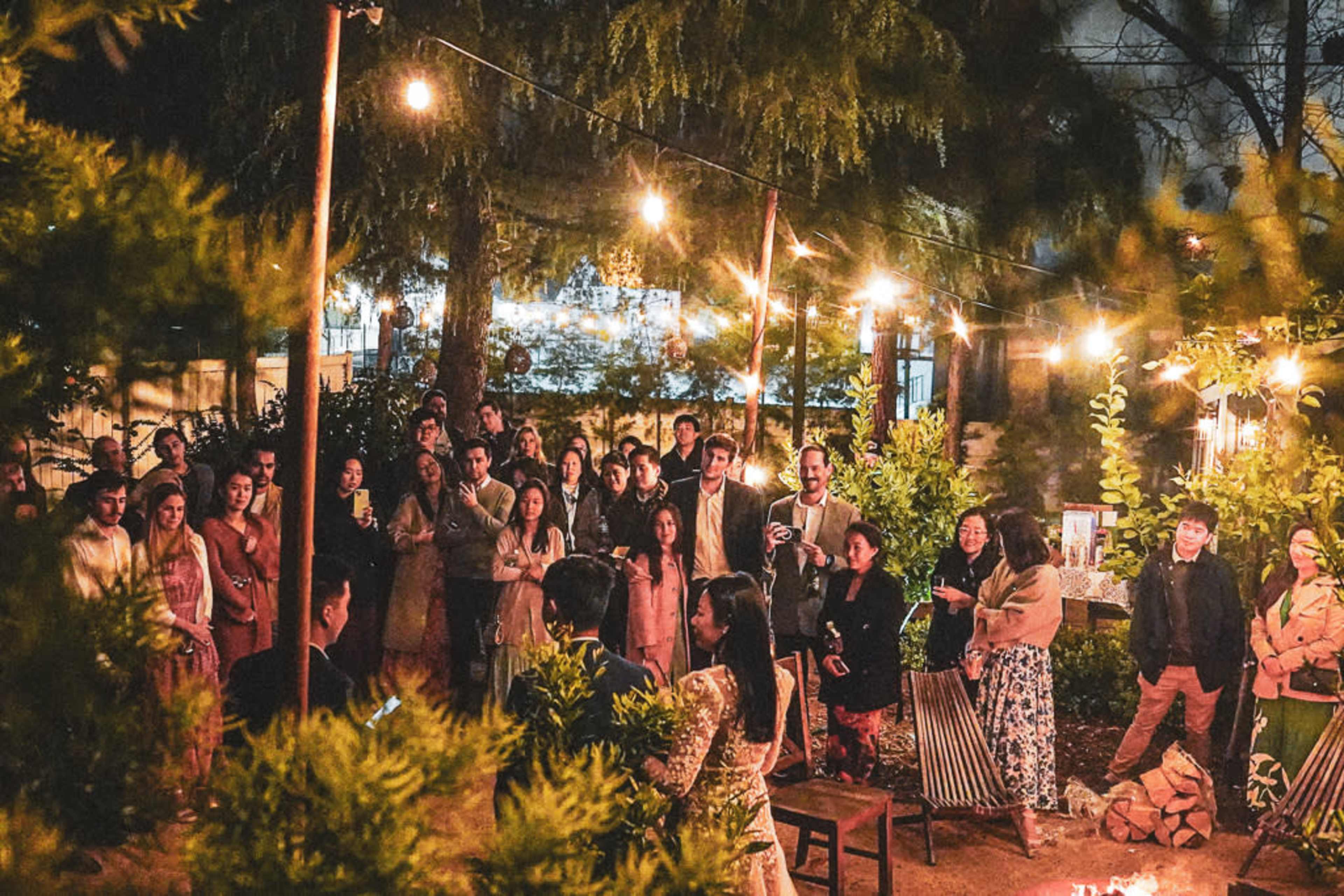 A crowd gathers around a woman speaking in a well-lit outdoor setting with string lights and trees.