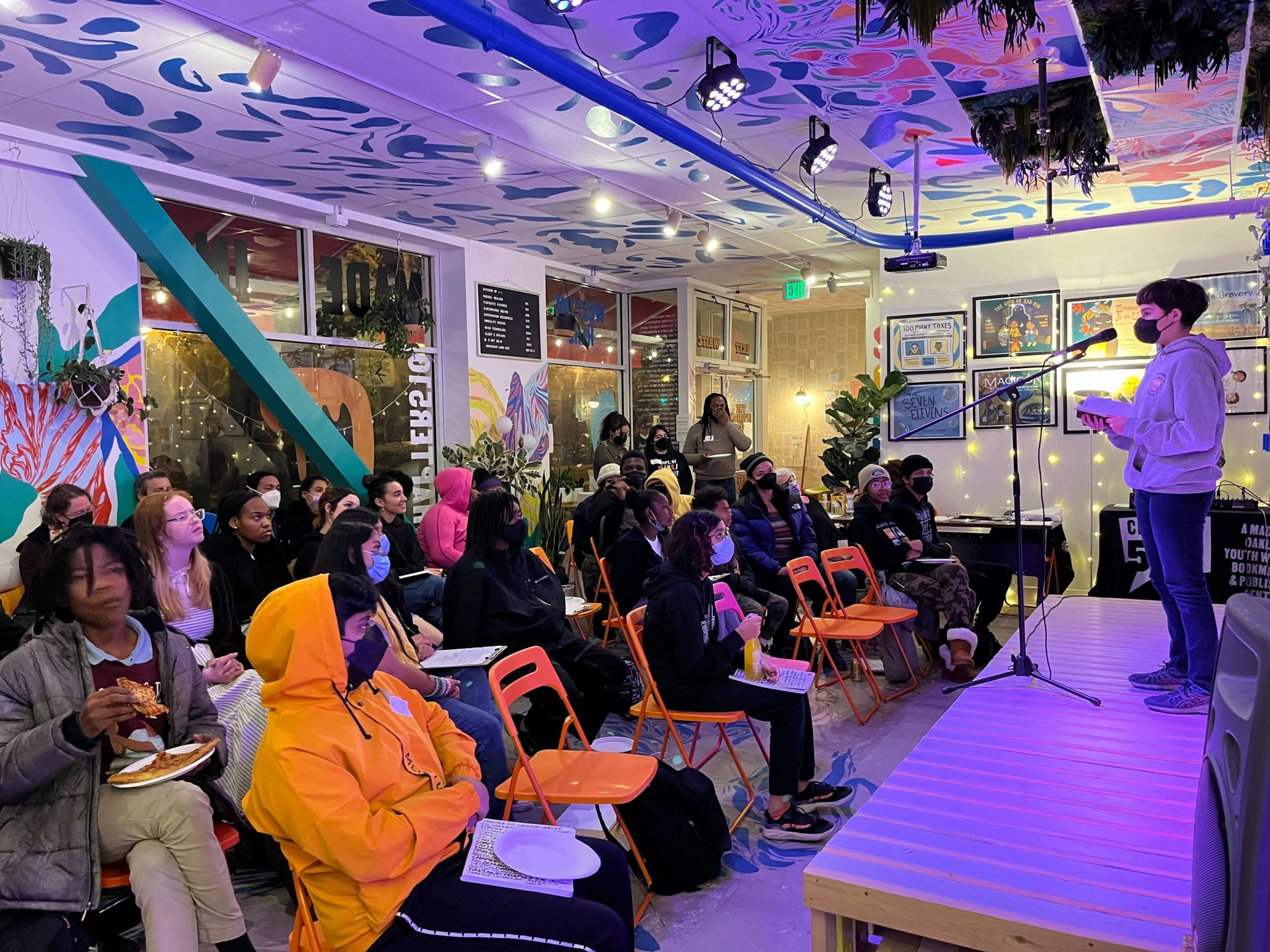A speaker stands on a platform in front of a seated audience in a brightly decorated room.