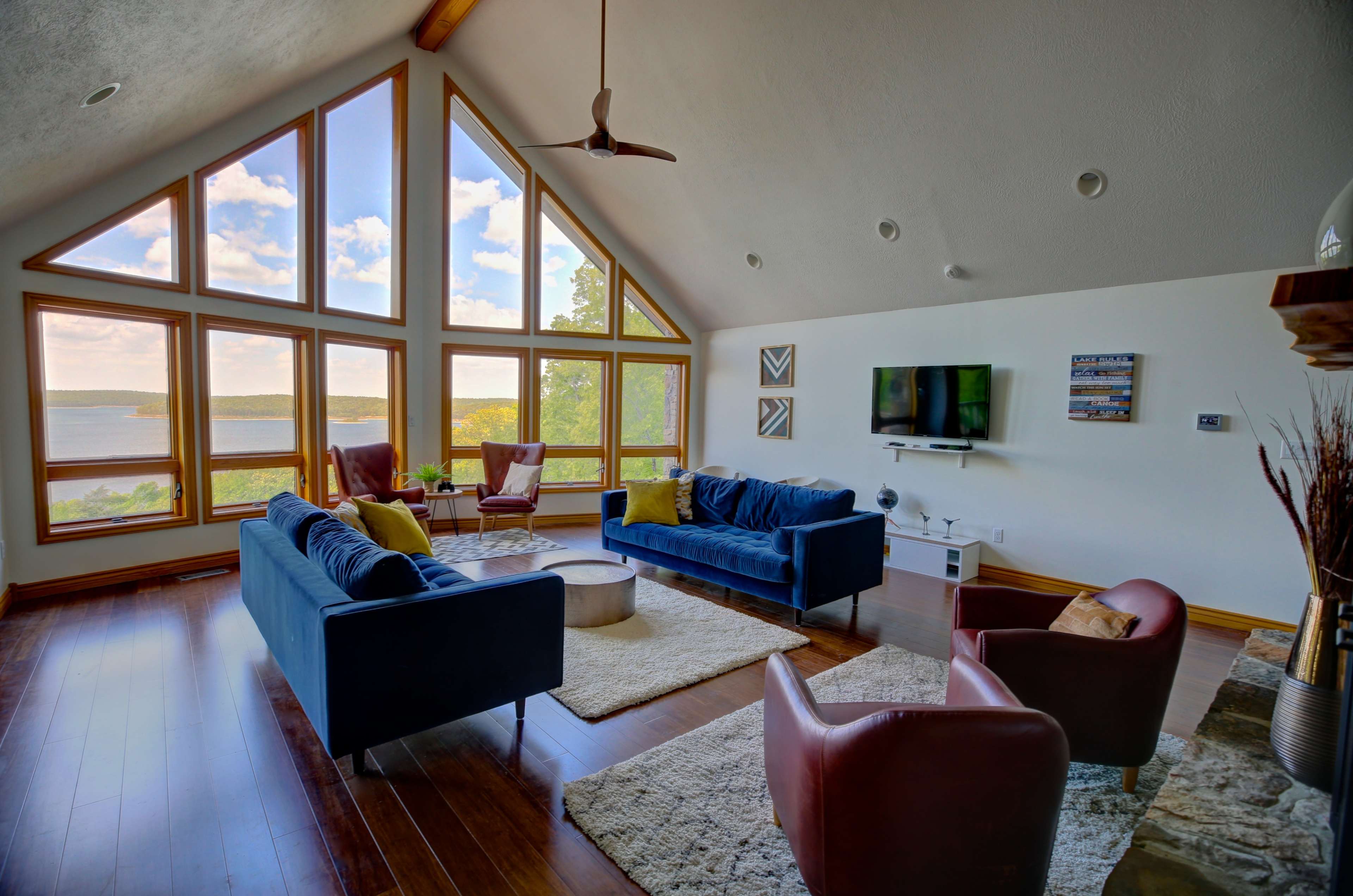 A spacious living room features large windows that offer a view of a lake and surrounding landscape, with two sofas, a coffee table, and a television.