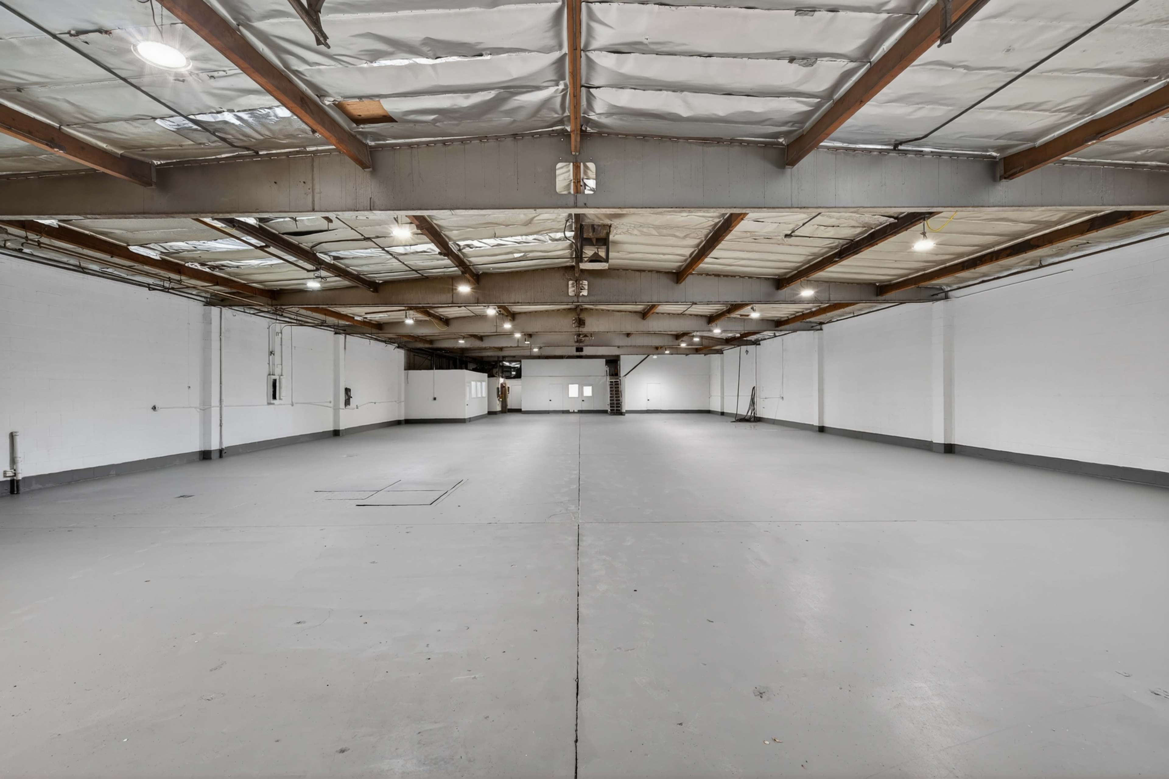The image shows a large, empty warehouse space with white walls, a flat gray floor, and exposed ceiling beams.